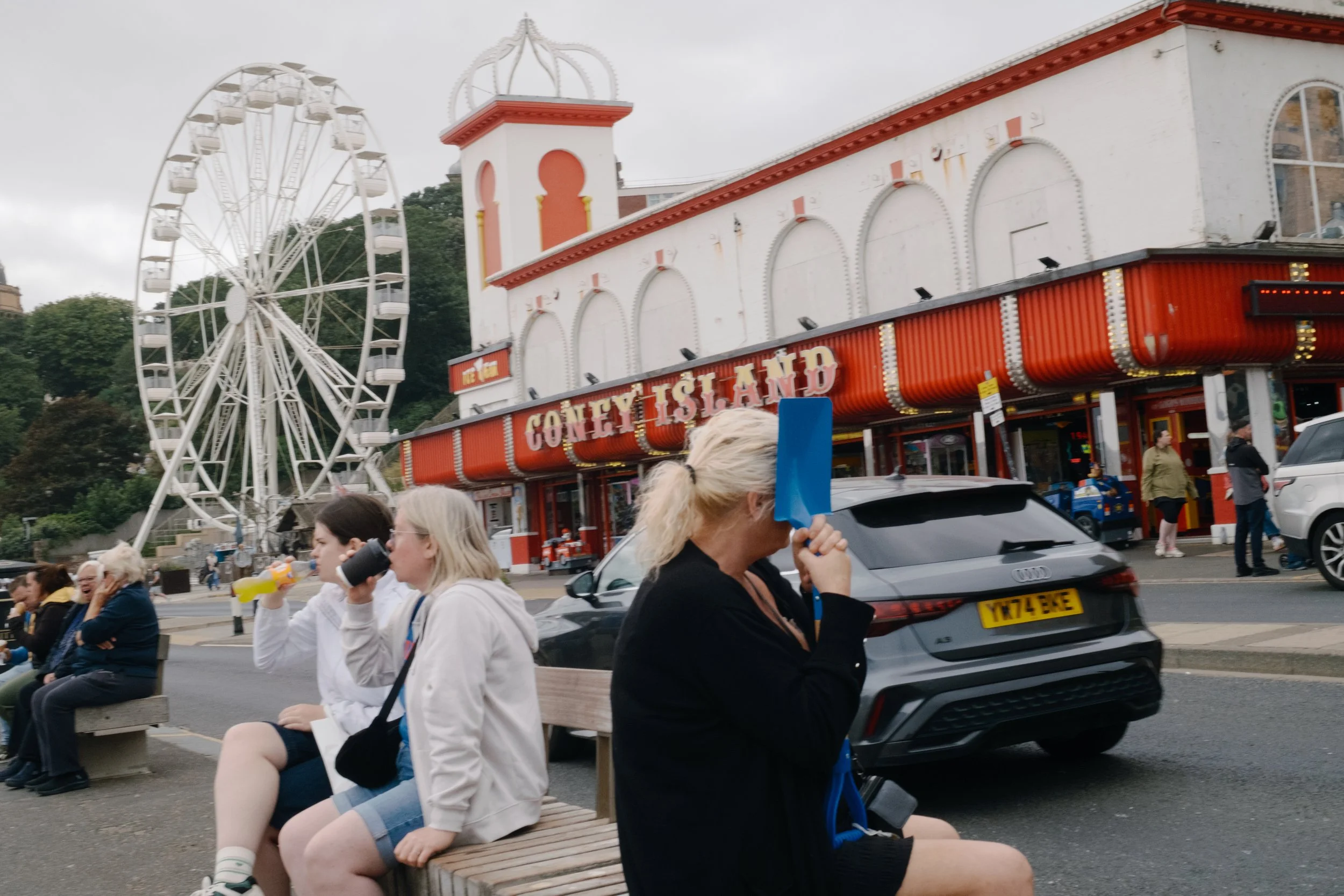 People sitting on benches at a crowd-filled outdoor amusement park, with a large white Ferris wheel and a red and white building labeled 'Coney Island' in the background.