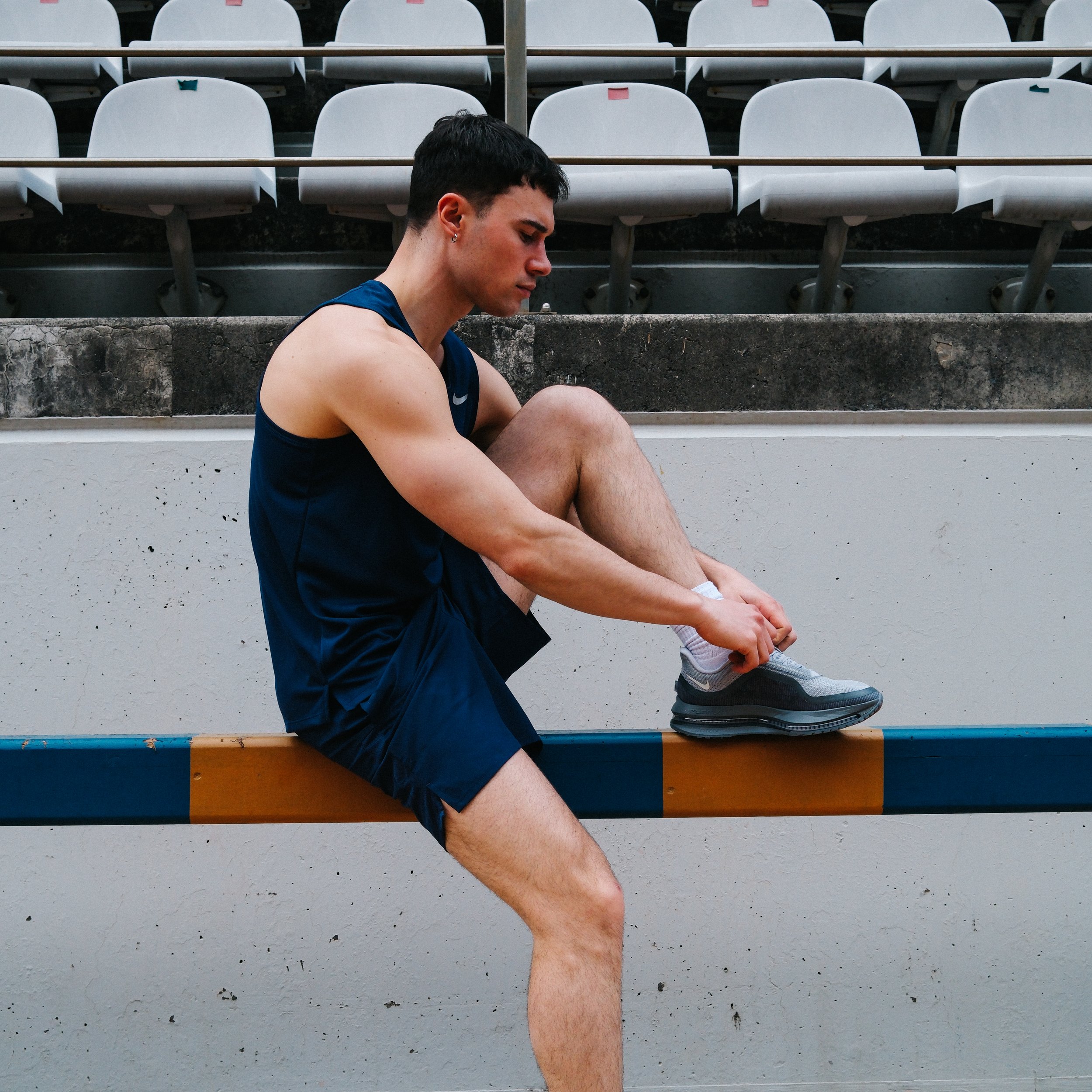 A man in a blue tank top is sitting on a bench with his head bowed and eyes closed, in front of empty white bleacher seats.