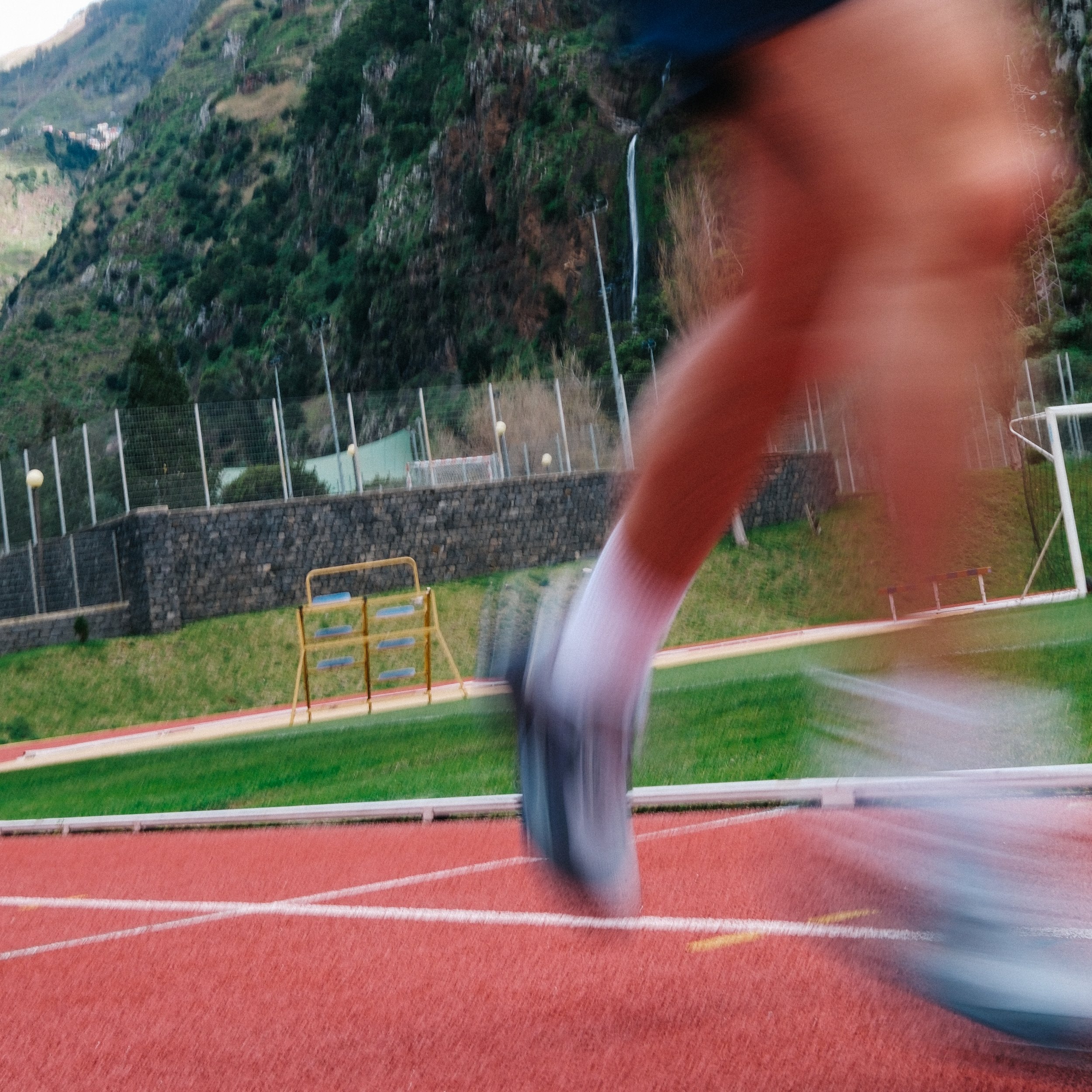 Outdoor sports track with a person running, a fenced area, and a hillside with trees and waterfalls in the background.