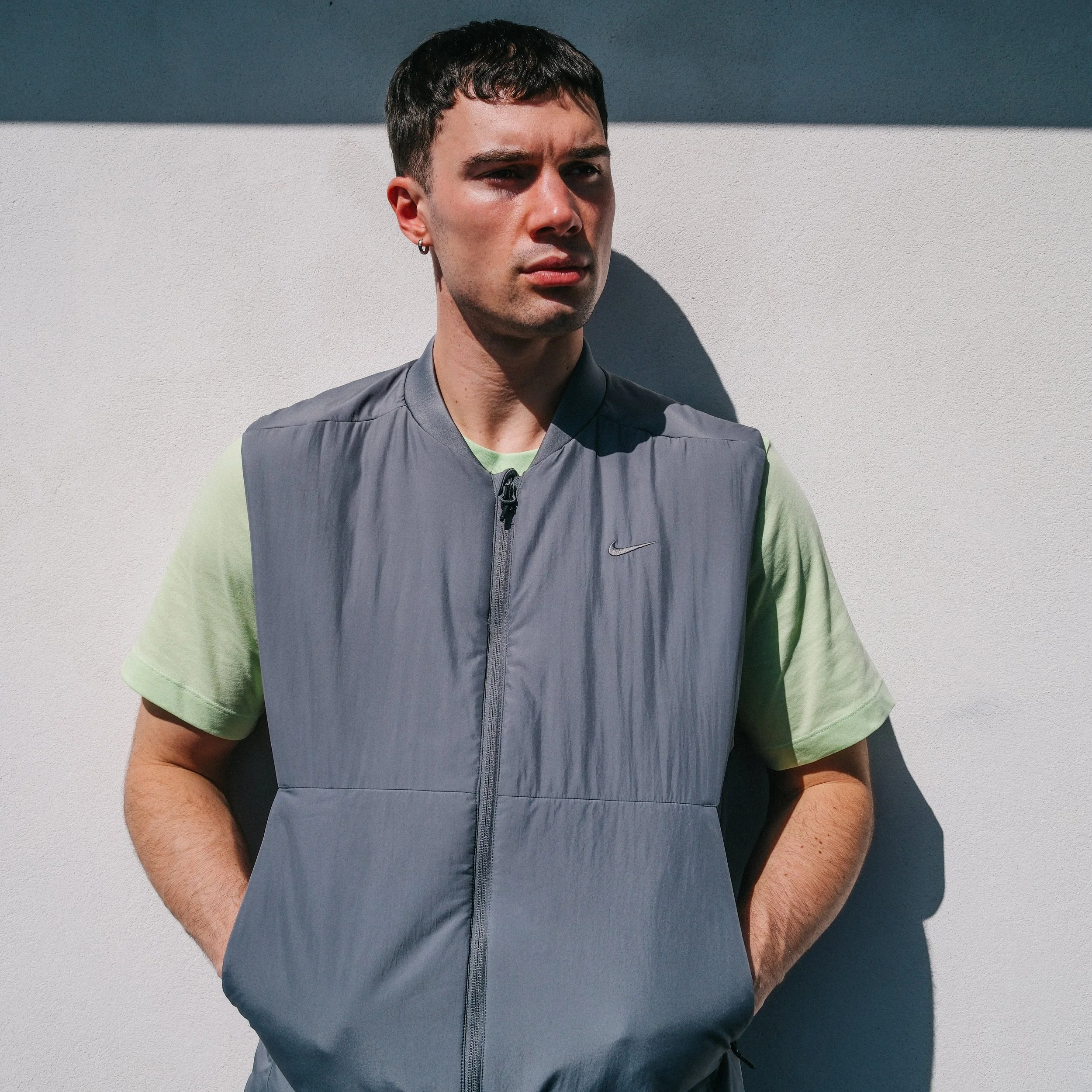 A young man with short dark hair, earrings, and a serious expression, wearing a light green t-shirt and a gray zip-up vest, stands against a white wall with a shadow cast from the sunlight.