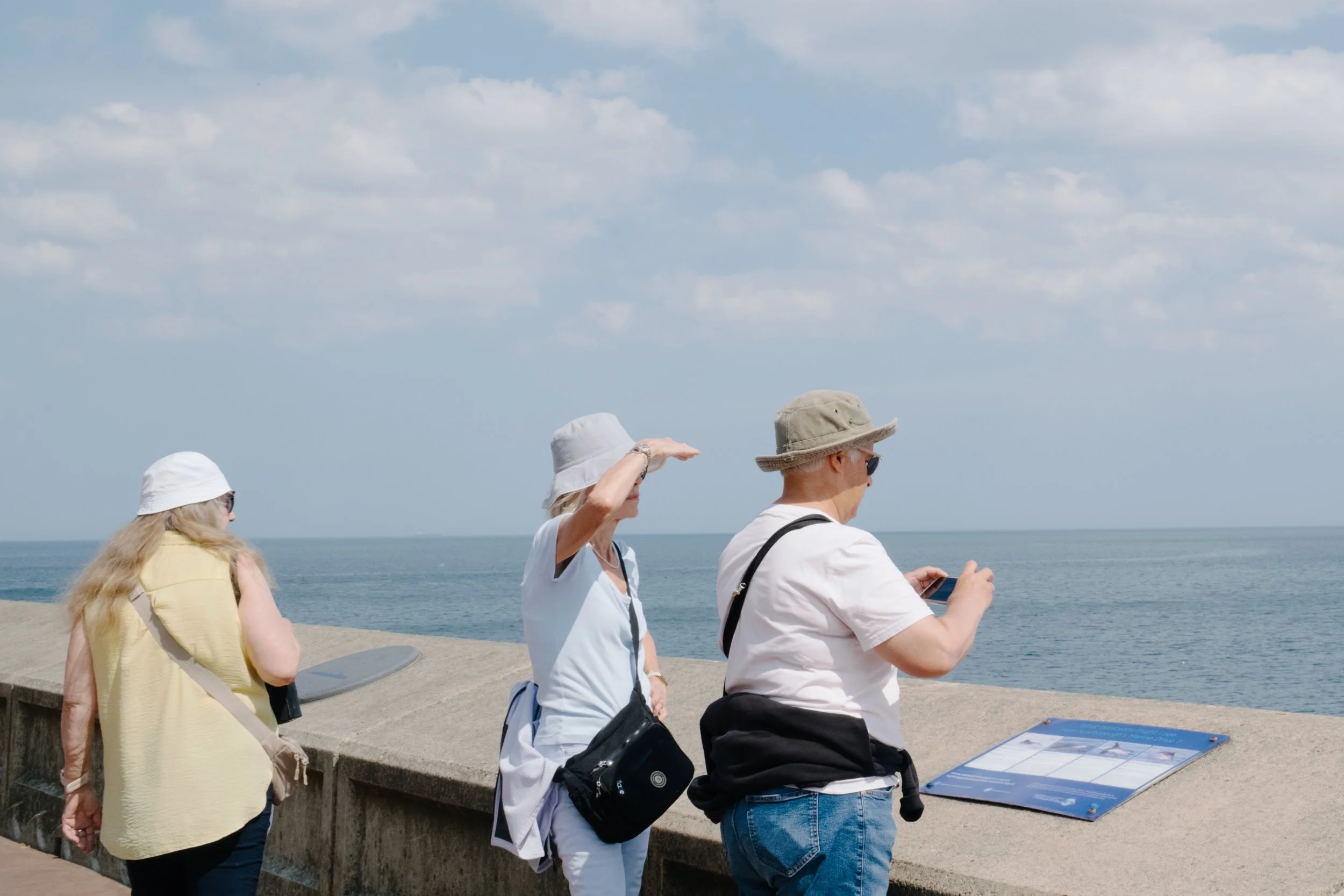 Four women walking along a beach promenade near the ocean, some wearing hats, one looking at her phone, under partly cloudy sky.