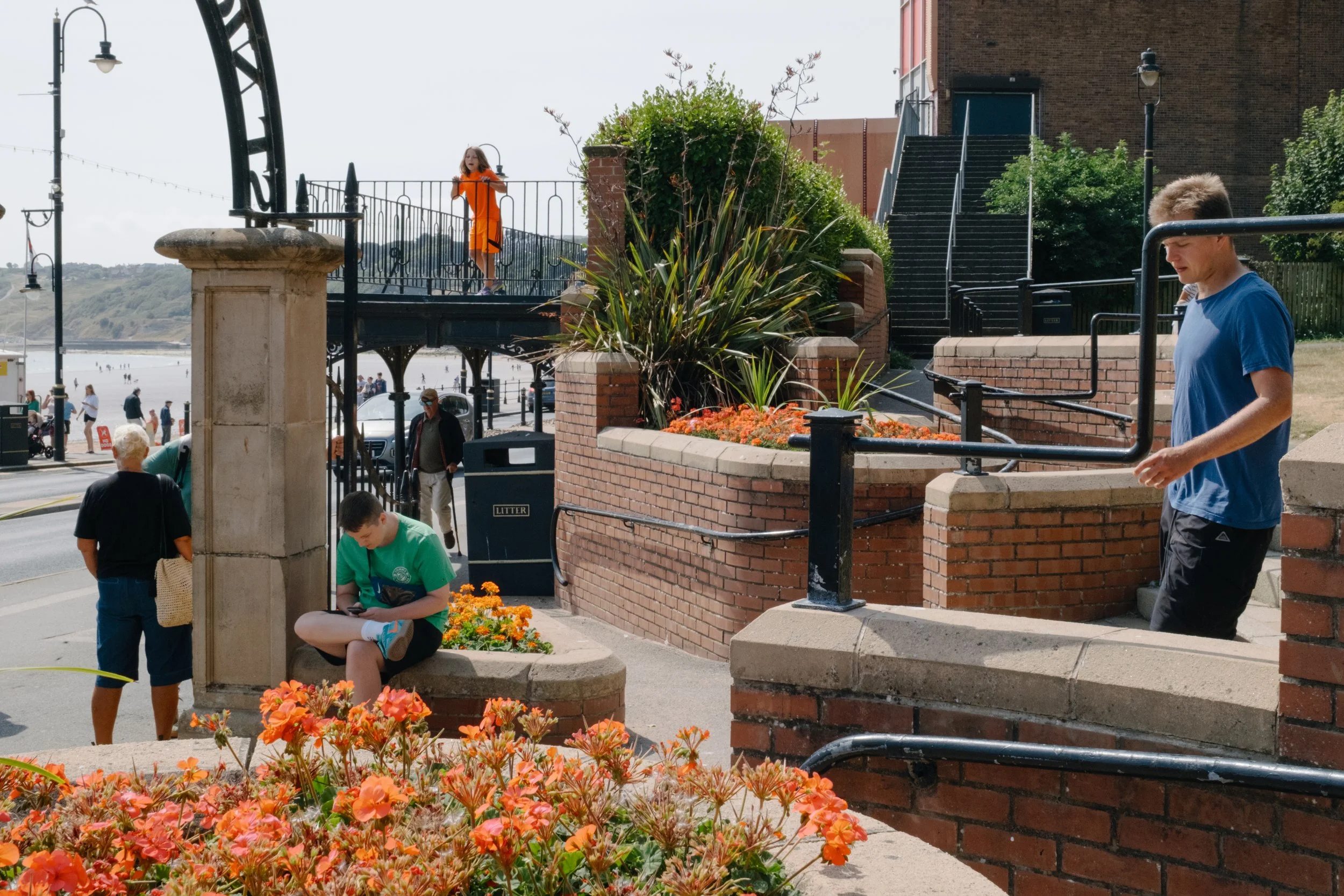 People on a sidewalk near orange and pink flowers, brick stairs, and a brick building, with some facing away and others looking at their phones, and a girl in an orange dress on a small bridge in the background.