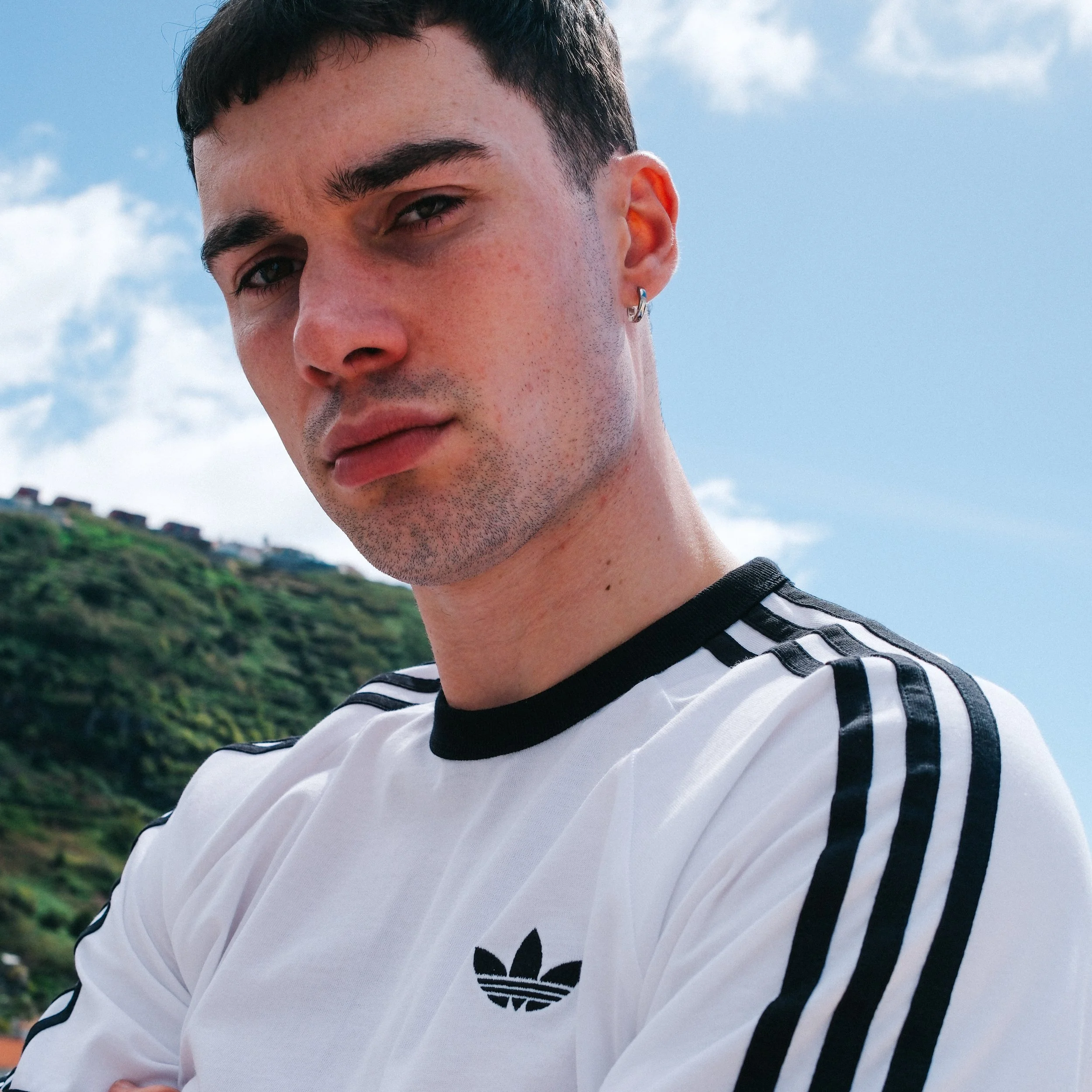 Close-up of a young man with dark hair, wearing a white Adidas shirt with black stripes, standing outdoors with a cloudy blue sky and green hillside in the background.