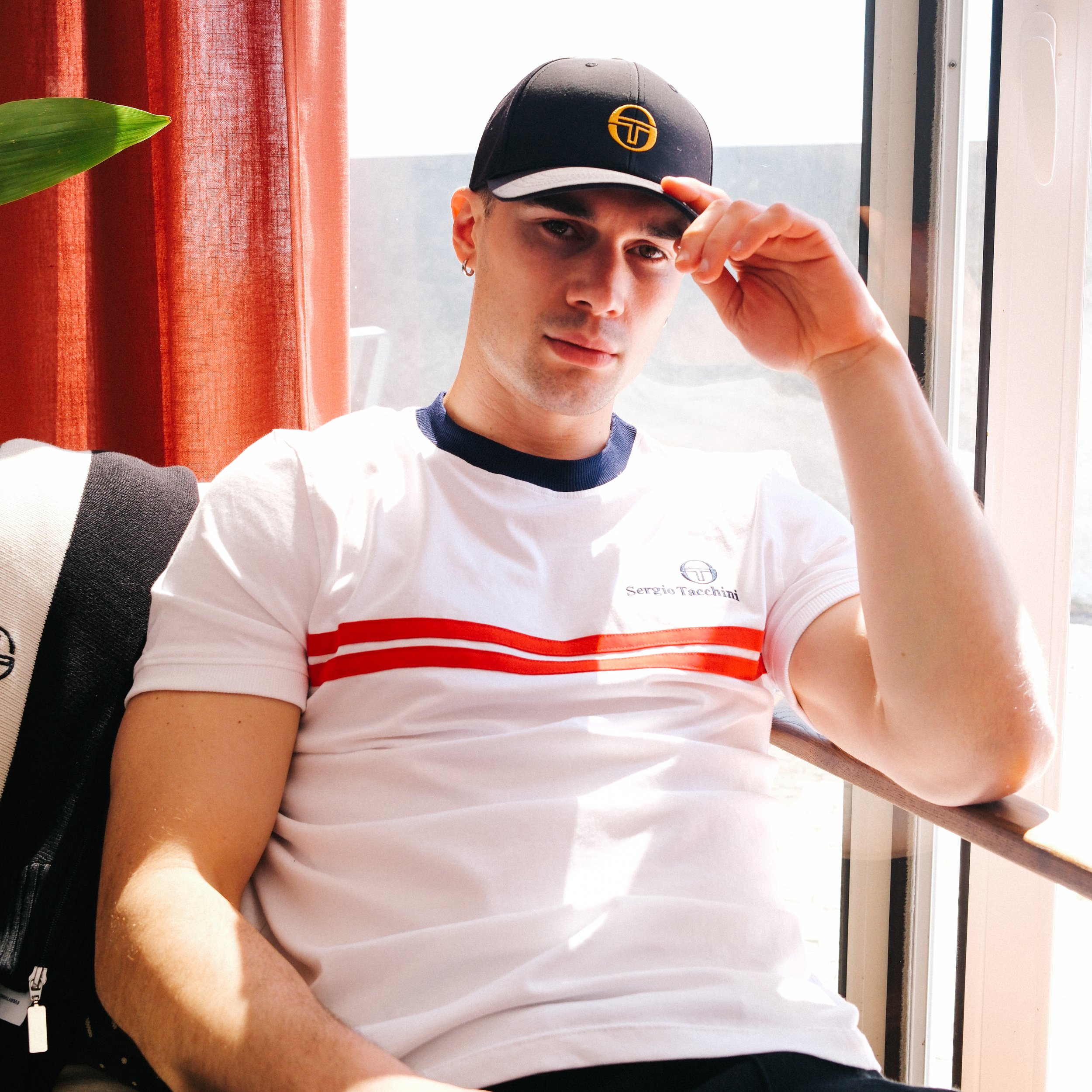 A young man wearing a black baseball cap with a yellow logo, a white T-shirt with red and blue stripes, and a small hoop earring, sitting by a window with red curtains and a green plant nearby, adjusting his cap.