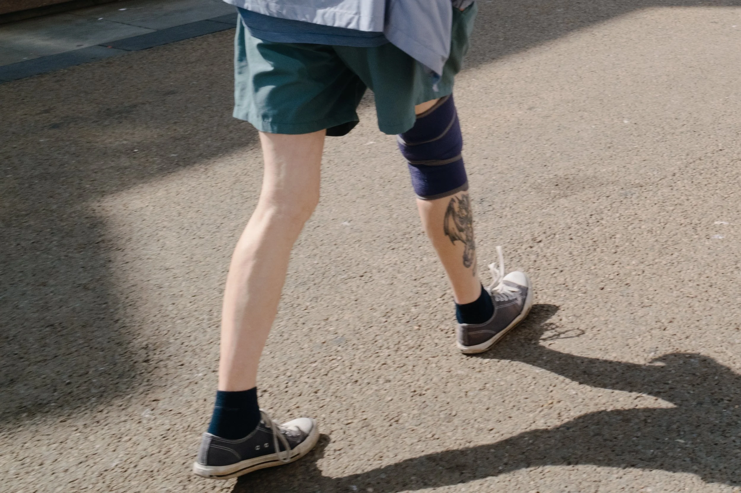 A person walking on a paved sidewalk, wearing dark shorts, a white shirt, and sneakers, with a tattoo on their calf.