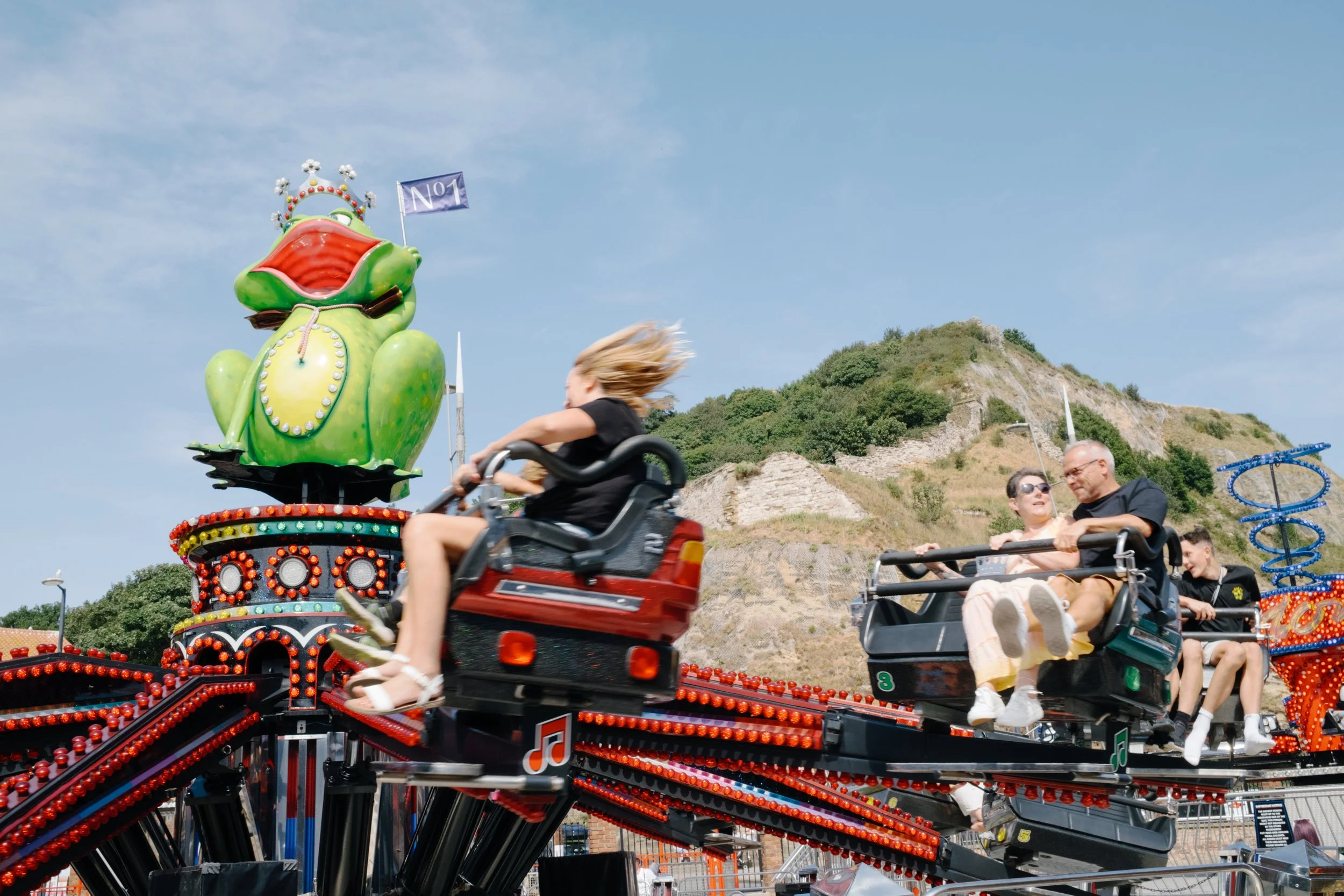 People riding a spinning amusement park ride shaped like a colorful frog, with hills and a clear blue sky in the background.