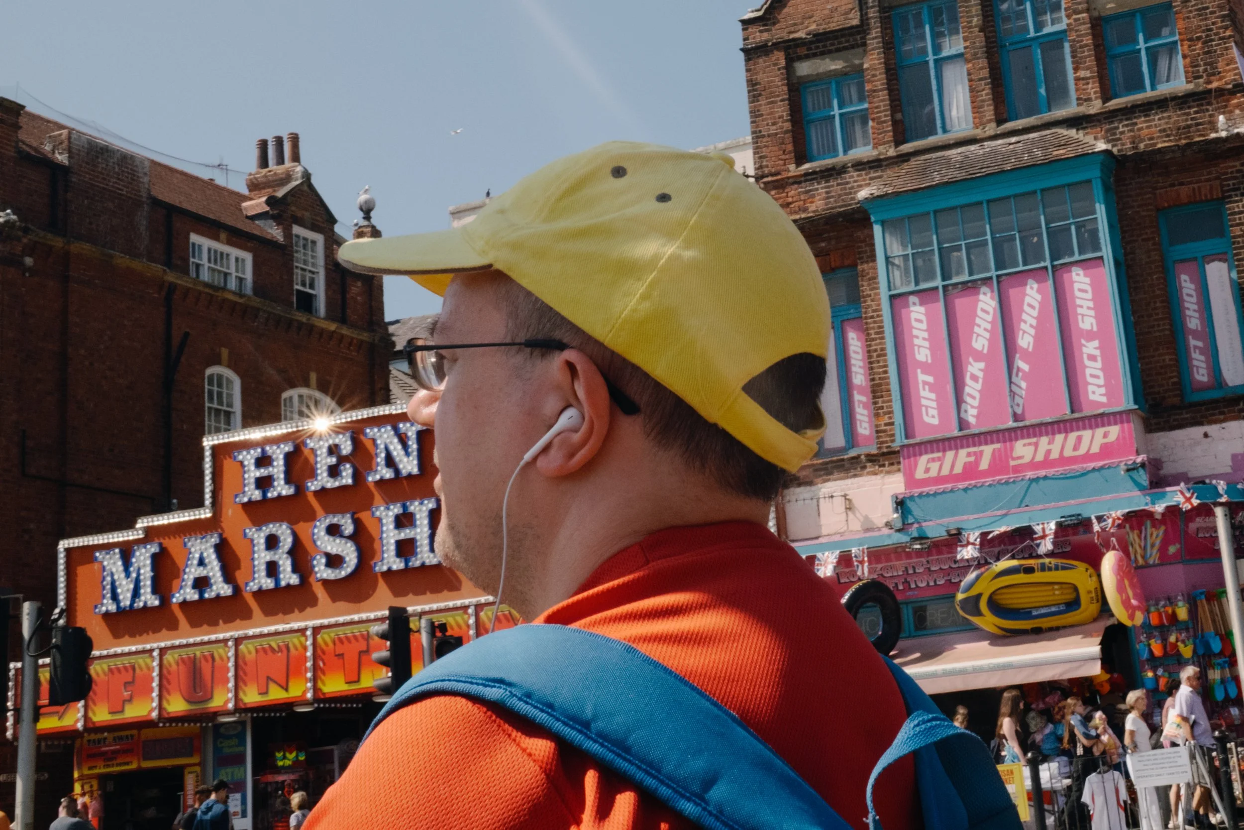 A person at a carnival wearing a yellow cap with a duck face design, glasses, and earphones, with colorful vendor booths and signs in the background.