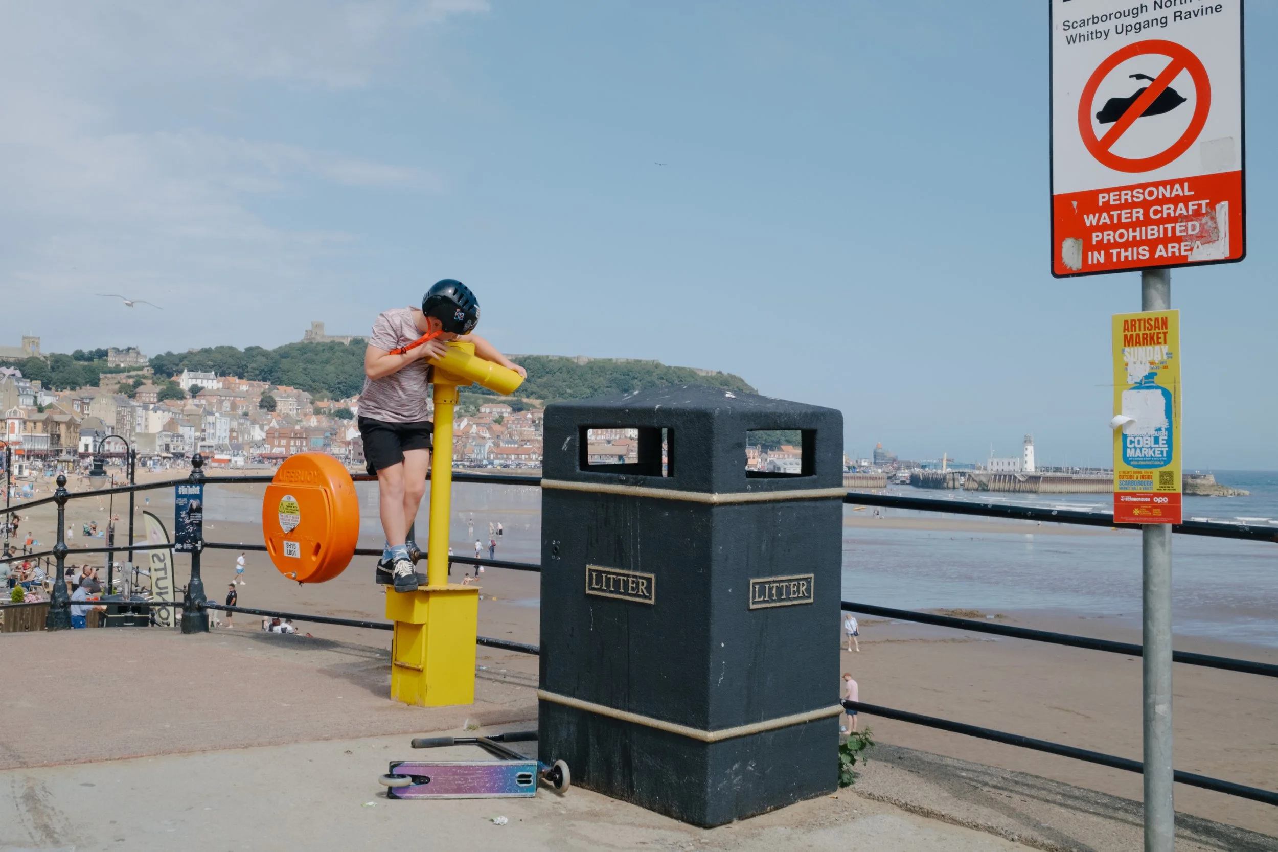 A child wearing a helmet and a striped shirt stands on a ledge, looking through a yellow binocular viewer, near a beach with a town and a lighthouse in the background. There are beachgoers below, a trash bin labeled 'LITTER', a sign prohibiting perso