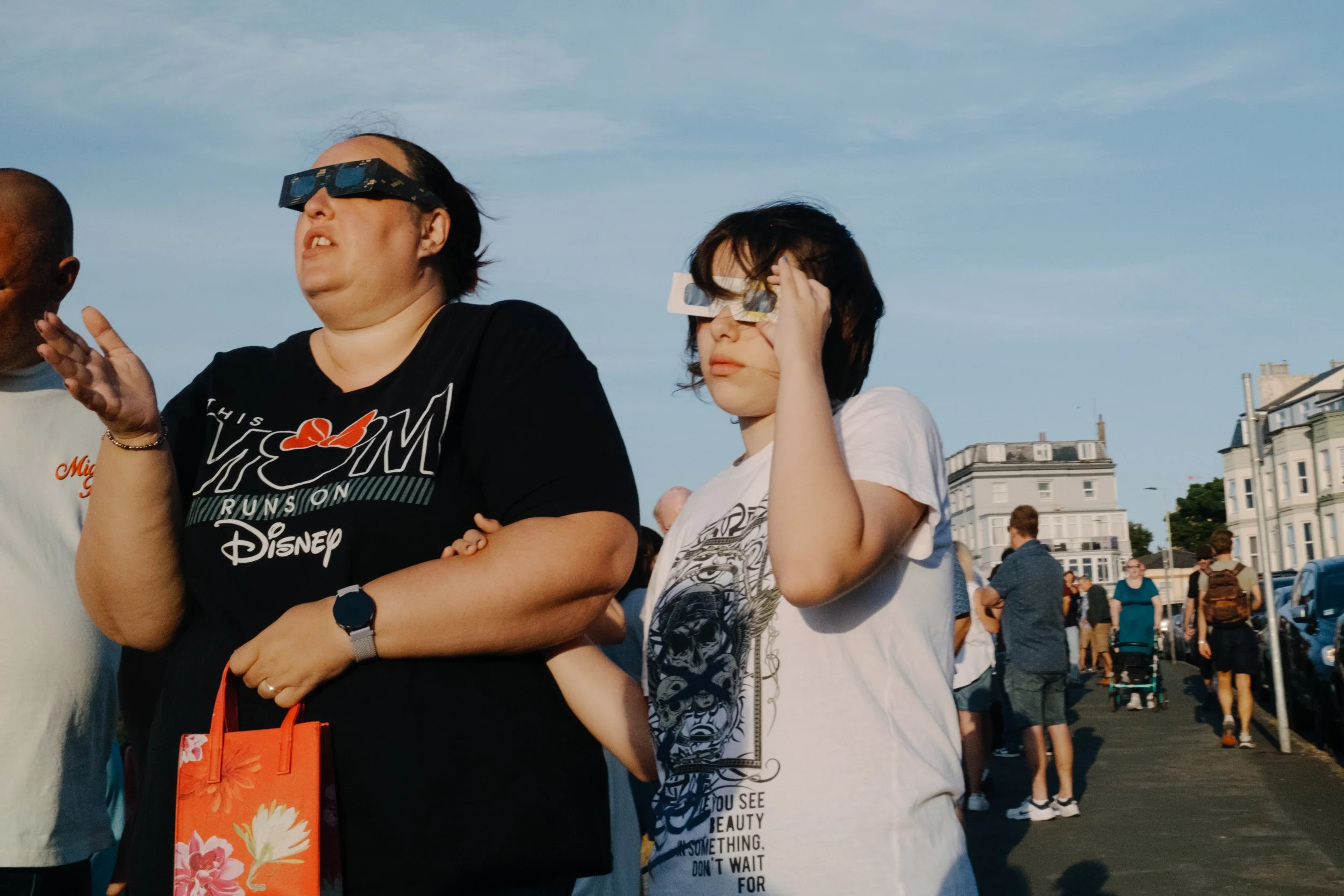 Two women standing outdoors among a crowd, with buildings in the background, appear to be at an event or festival. One woman is wearing NASA-themed glasses and a white t-shirt, with her hand near her face. The other woman is wearing a black T-shirt w