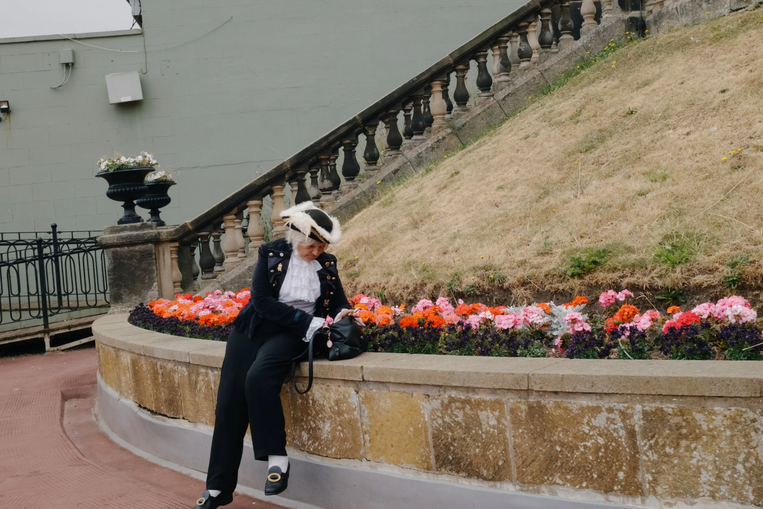 Woman dressed in historical costume sitting on a stone bench, surrounded by colorful flowers, near a staircase with balustrades, in an outdoor setting.