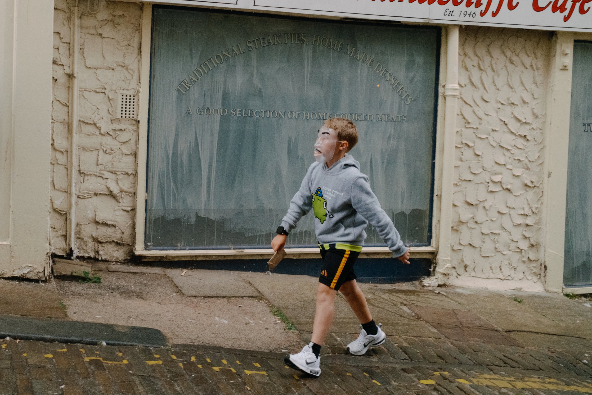 A young boy wearing a gray hoodie with a green cartoon character on it and black shorts runs along a sidewalk in front of a storefront window. The window has writing that promotes homemade cooked meats and traditional steak pies. The building has a t