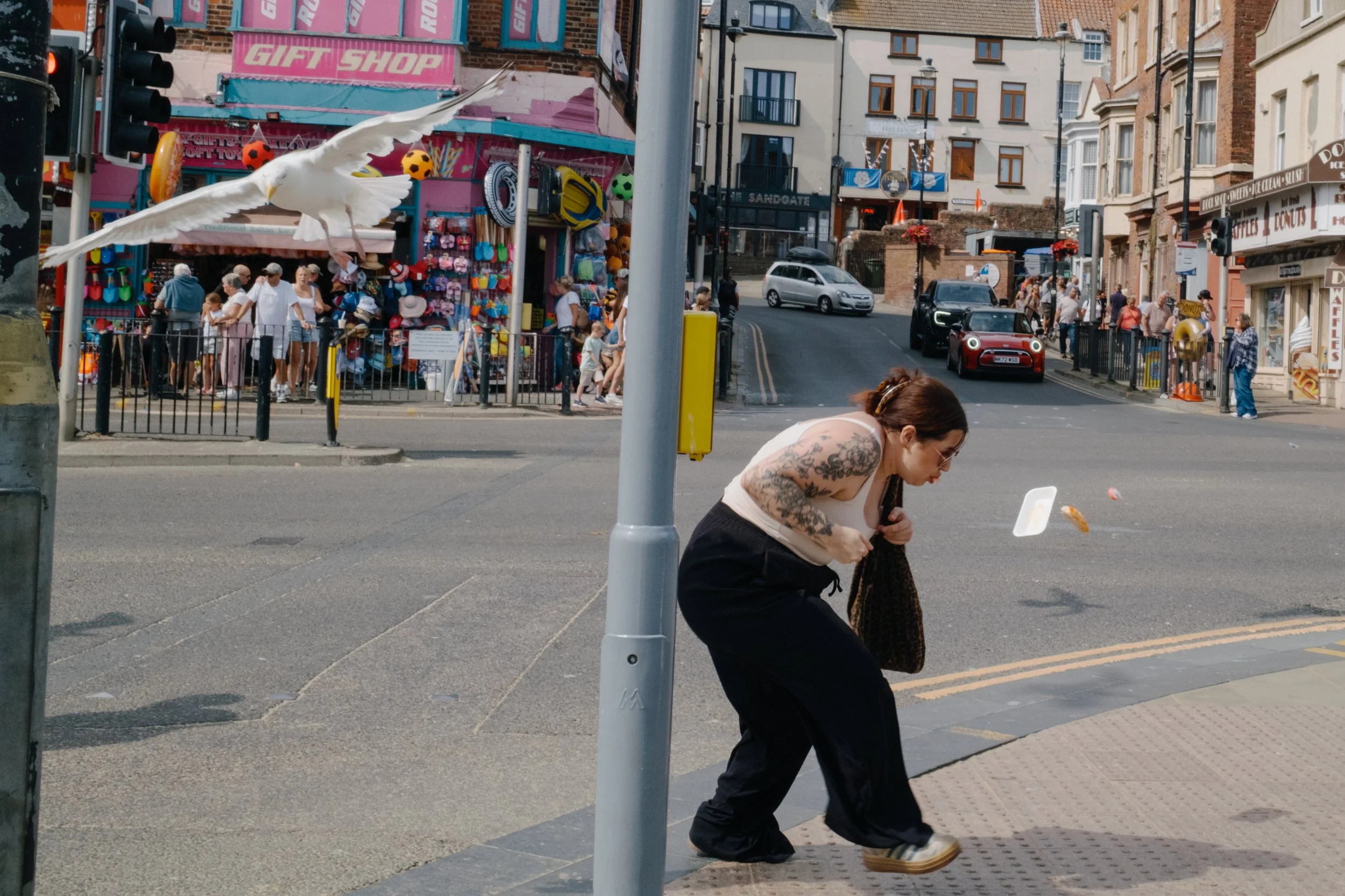 A busy street scene with buildings, shops, and cars. People are walking on the sidewalk, and a woman with tattoos is bending down near the curb. A large seagull is flying in the foreground. There is a gift shop with pink signage and multicolored item
