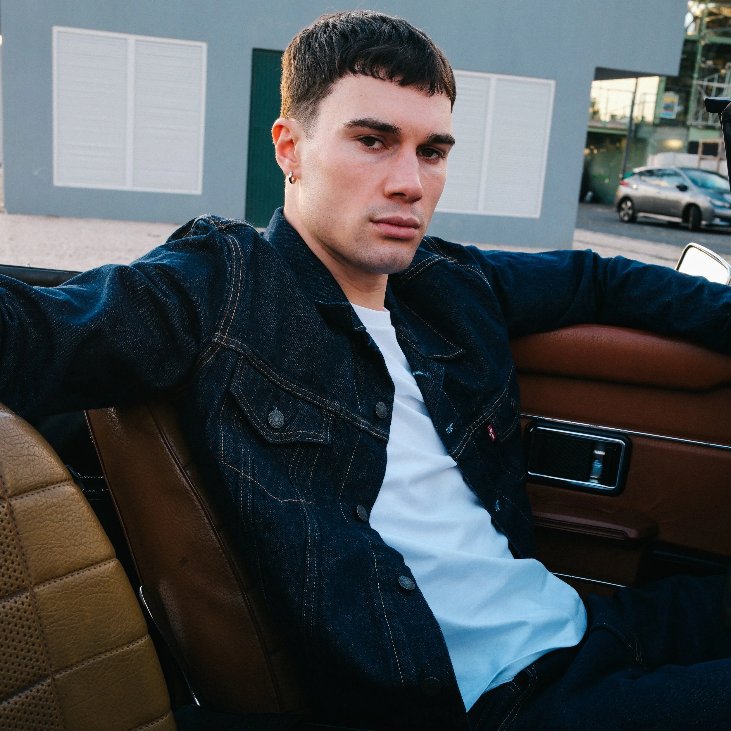 A young man with short dark hair and pierced ears sitting in a vintage car, wearing a dark denim jacket and white shirt, outside in front of a modern building with parked cars in the background.