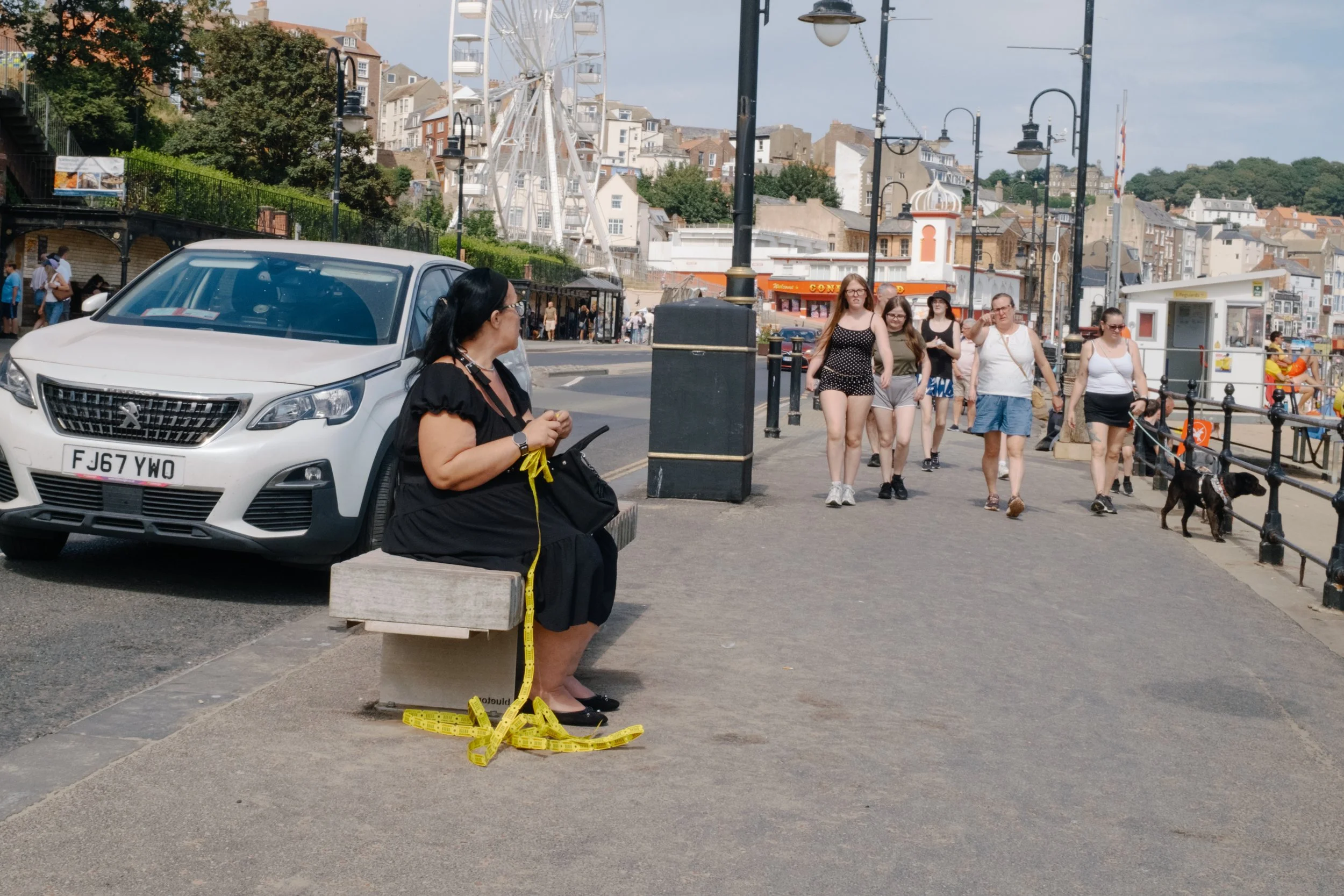A woman sitting on a bench on a busy street, with a cardboard sign around her neck and yellow measuring tape on the ground in front of her, while a group of people walk past nearby.