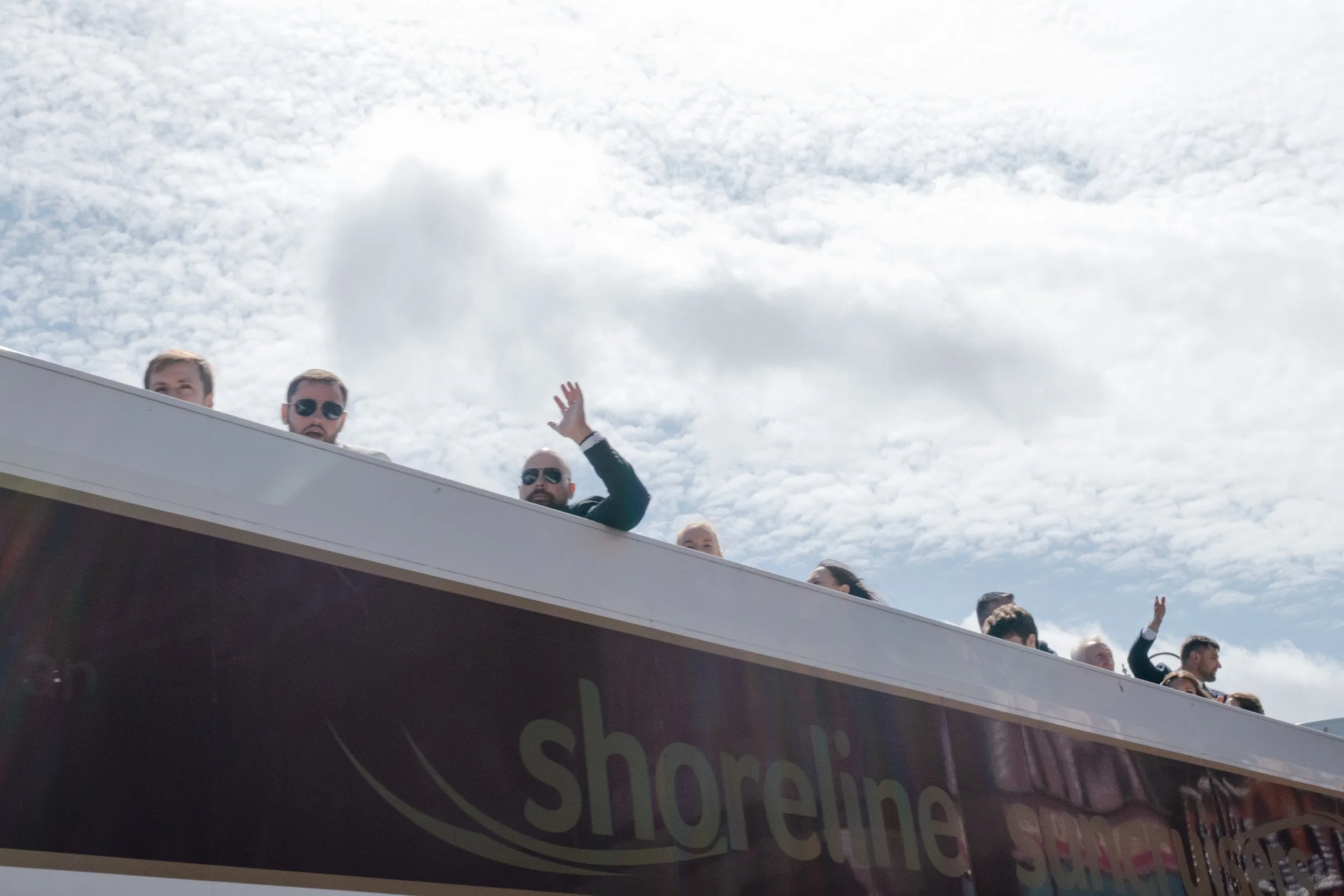 Group of people on a boat or platform, some wearing sunglasses, waving or looking in various directions against a partly cloudy sky.