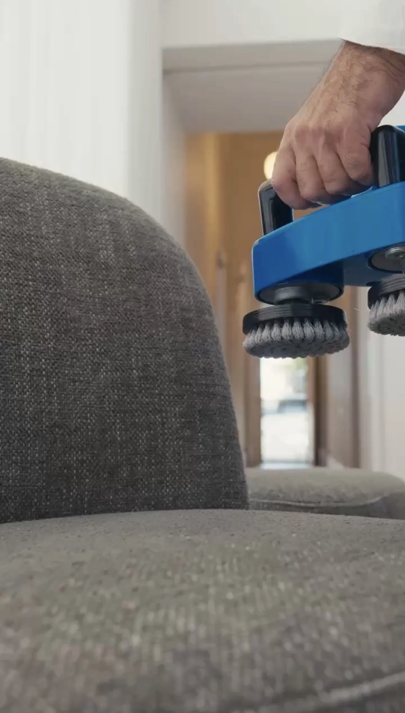 Close-up of a person using a handheld vacuum cleaner to clean the gray carpet of a sofa. The scene is indoors with a wooden door and a window in the background.