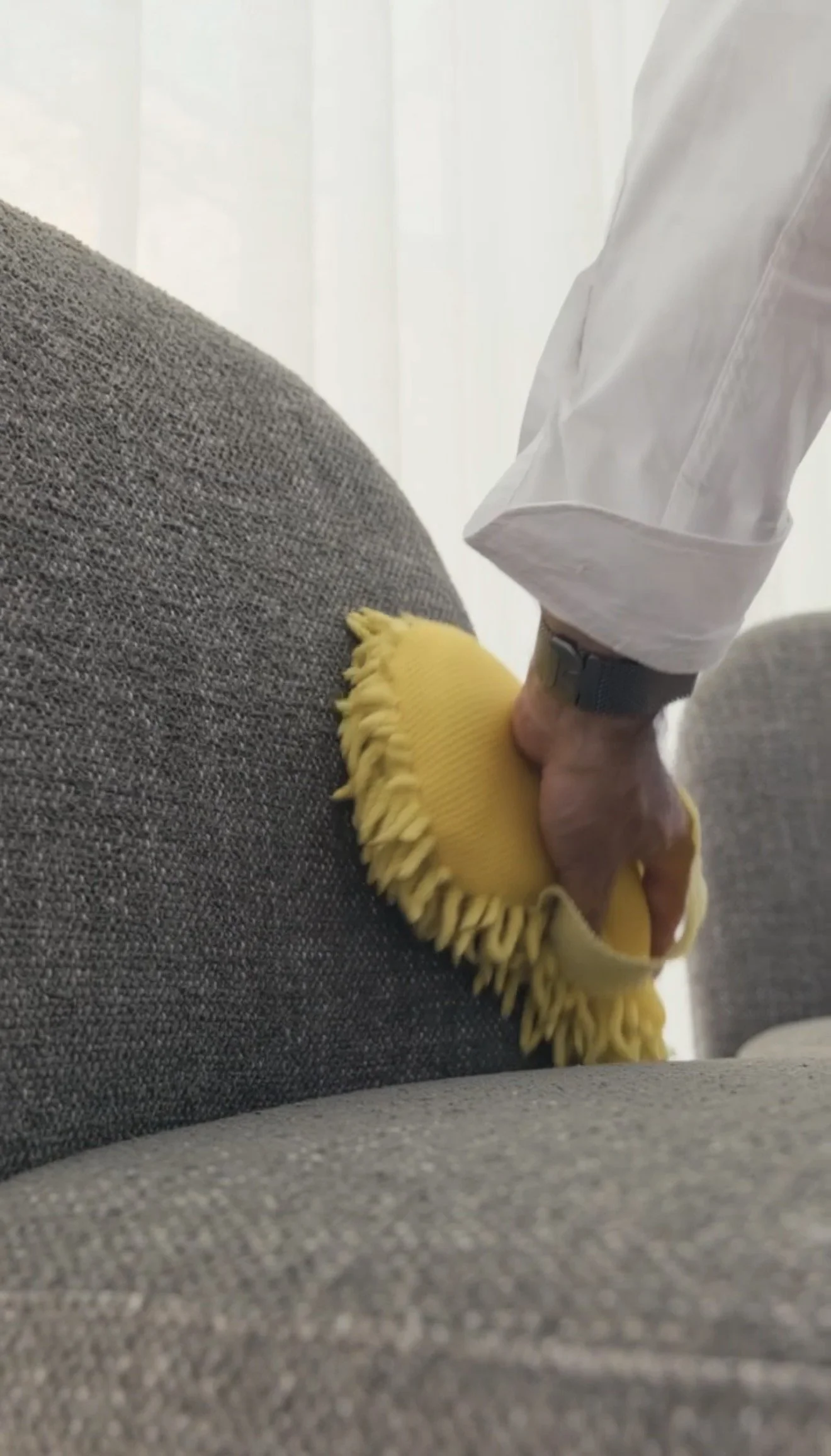 Person cleaning a gray textured sofa with a yellow microfiber duster.
