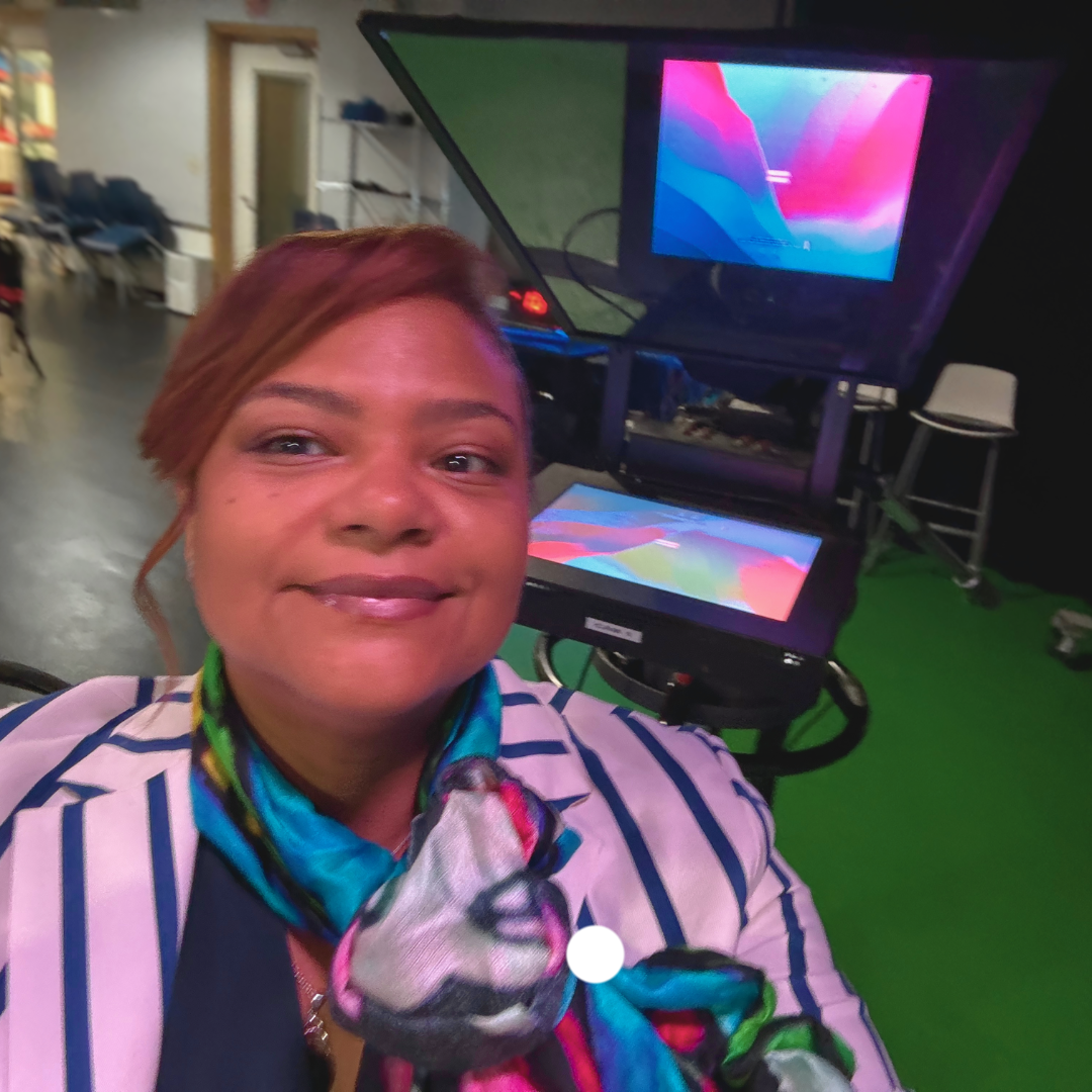 A woman taking a selfie with a green screen and large monitor in the background. She is wearing a striped blazer and a colorful scarf.