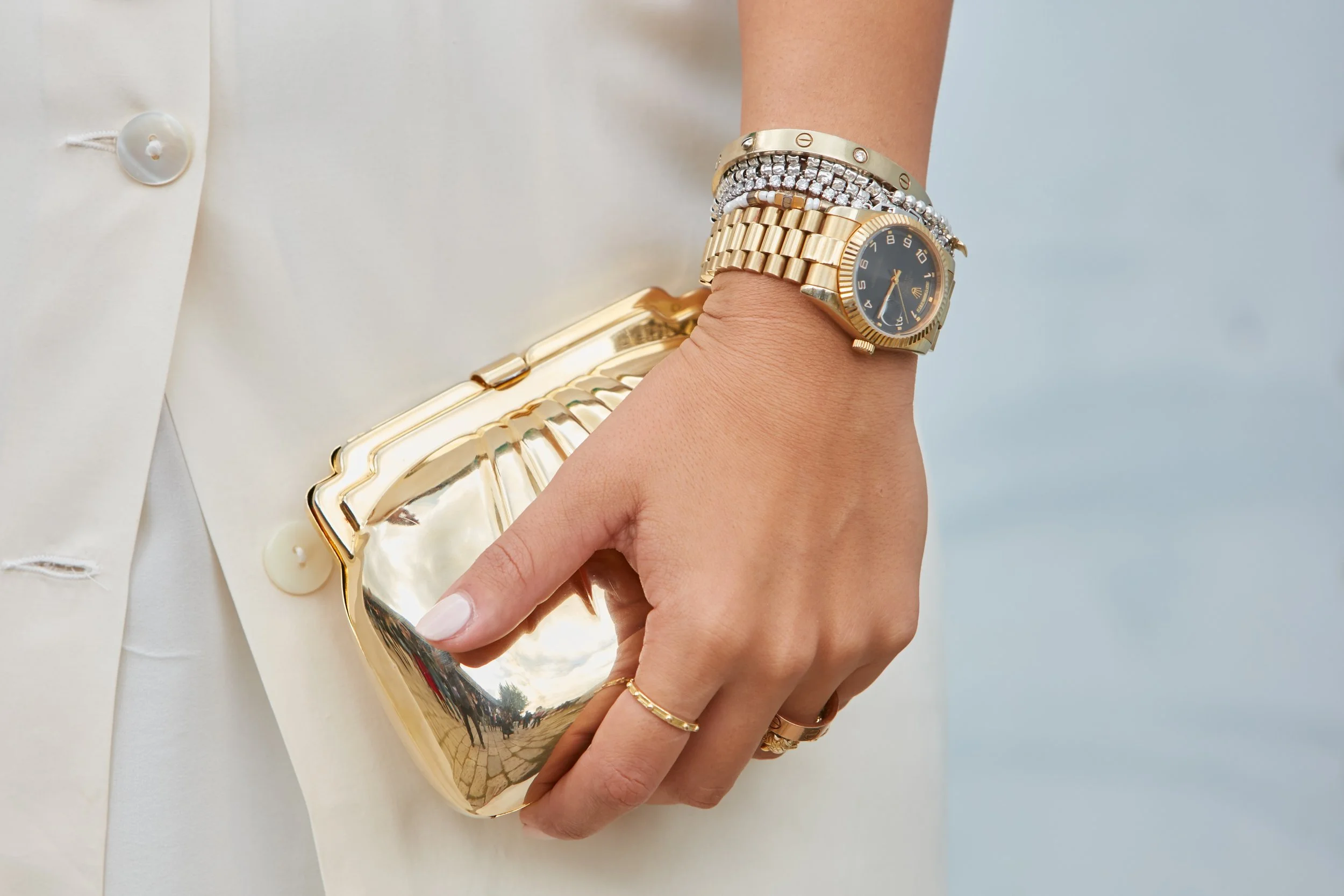A woman's hand holding a shiny gold clutch purse, wearing multiple gold and diamond bracelets and a gold watch, with part of a cream-colored jacket in the background.