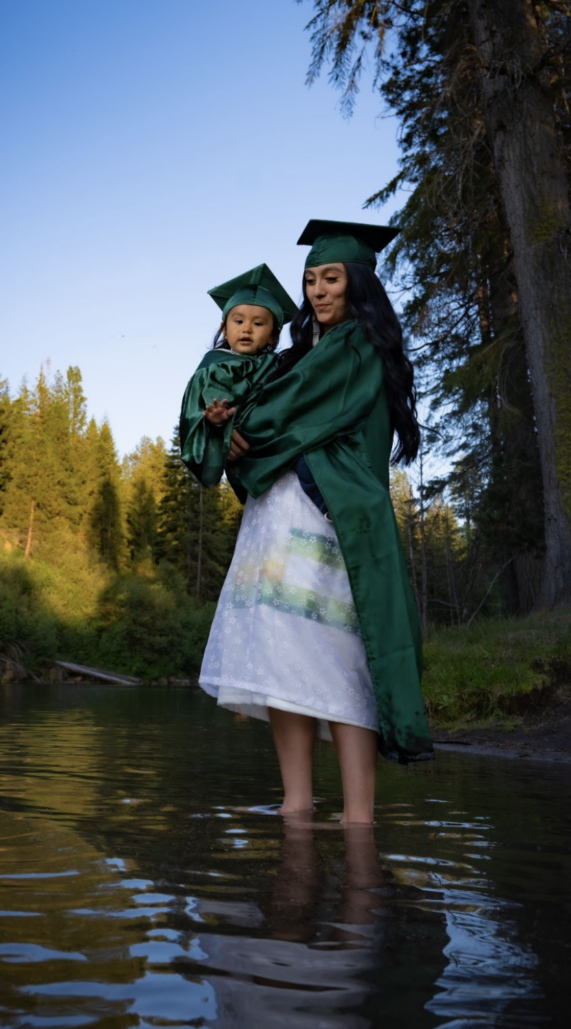 A woman and a young girl in green graduation gowns and caps stand in a shallow river surrounded by trees during daylight.