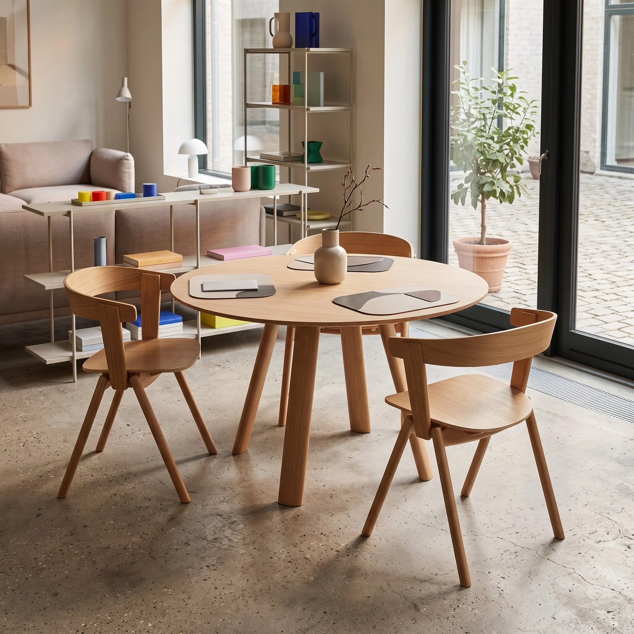 Modern living room with a round wooden table surrounded by wooden chairs, a beige sofa, a white shelving unit with decorative objects, large windows with a view of an outdoor patio, and a small potted tree.