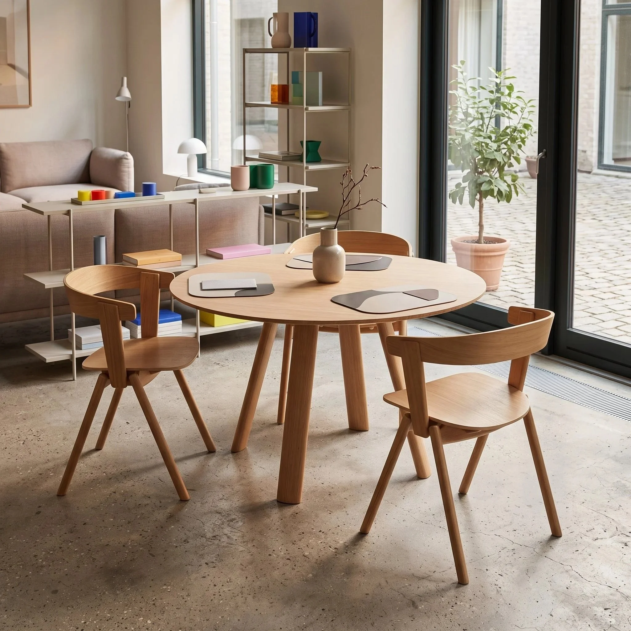 Interior of a modern living space with wooden dining table and chairs, decorative vases and books, large floor-to-ceiling windows with a view of outdoor potted plants.