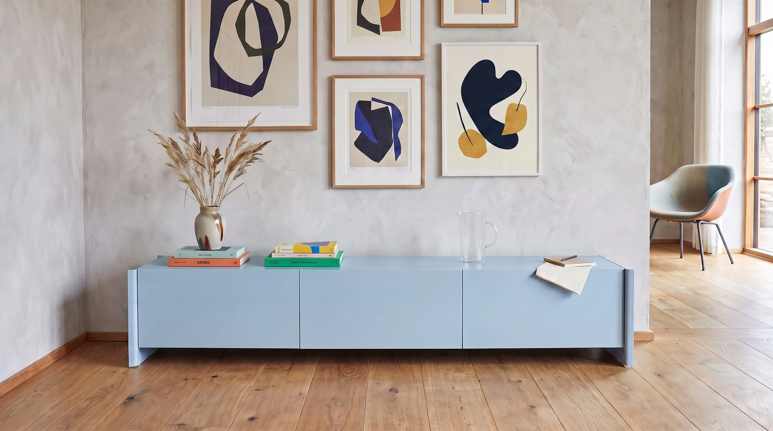 A light blue sideboard with a vase of dried pampas grass, several stacked books, a glass pitcher, and some notebooks on top. Behind it, the wall displays framed abstract artwork. To the right, a chair with a curved, two-tone seat is placed by a large