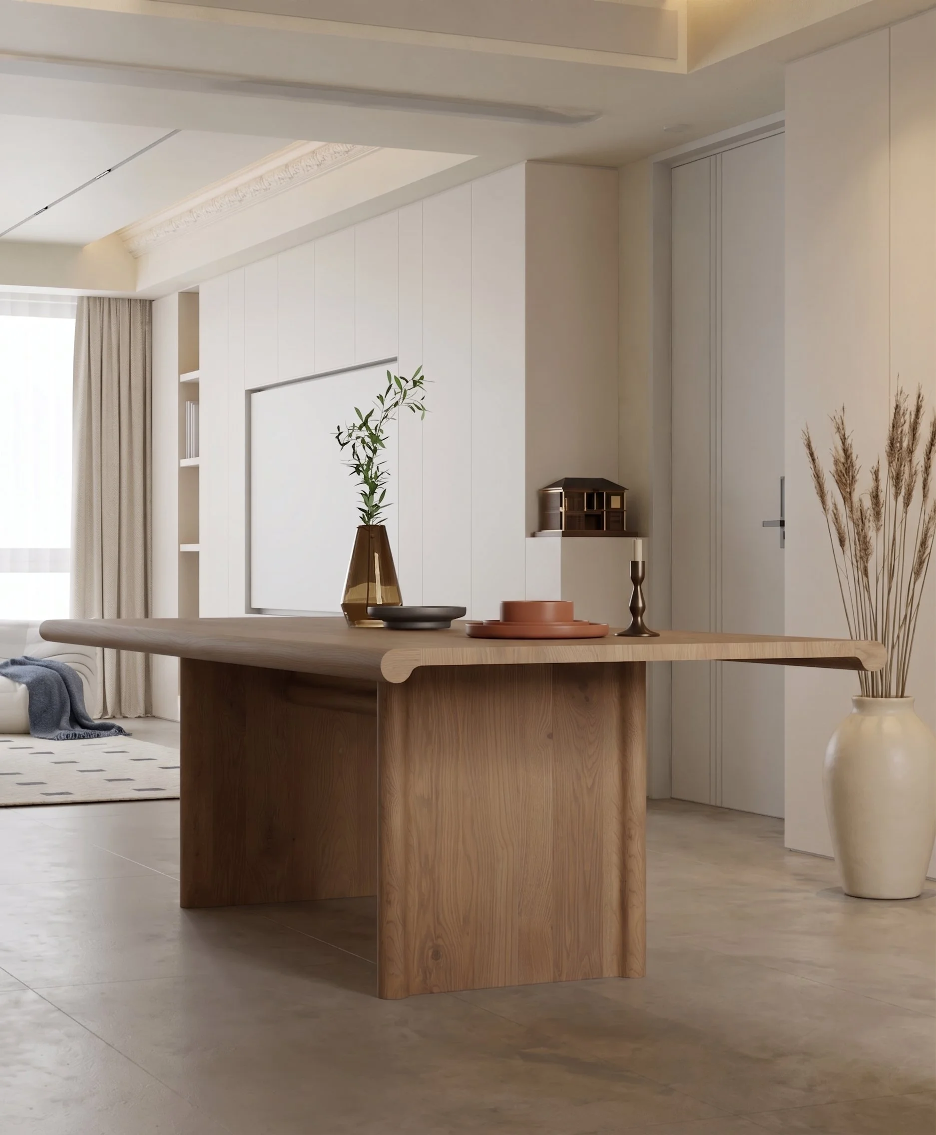 A modern living room with a large wooden table in the foreground, resting on a light-colored floor. Decorative items, including vases and a bowl, are arranged on the table. The room has a built-in white wall with minimalist shelving and a tall, narro