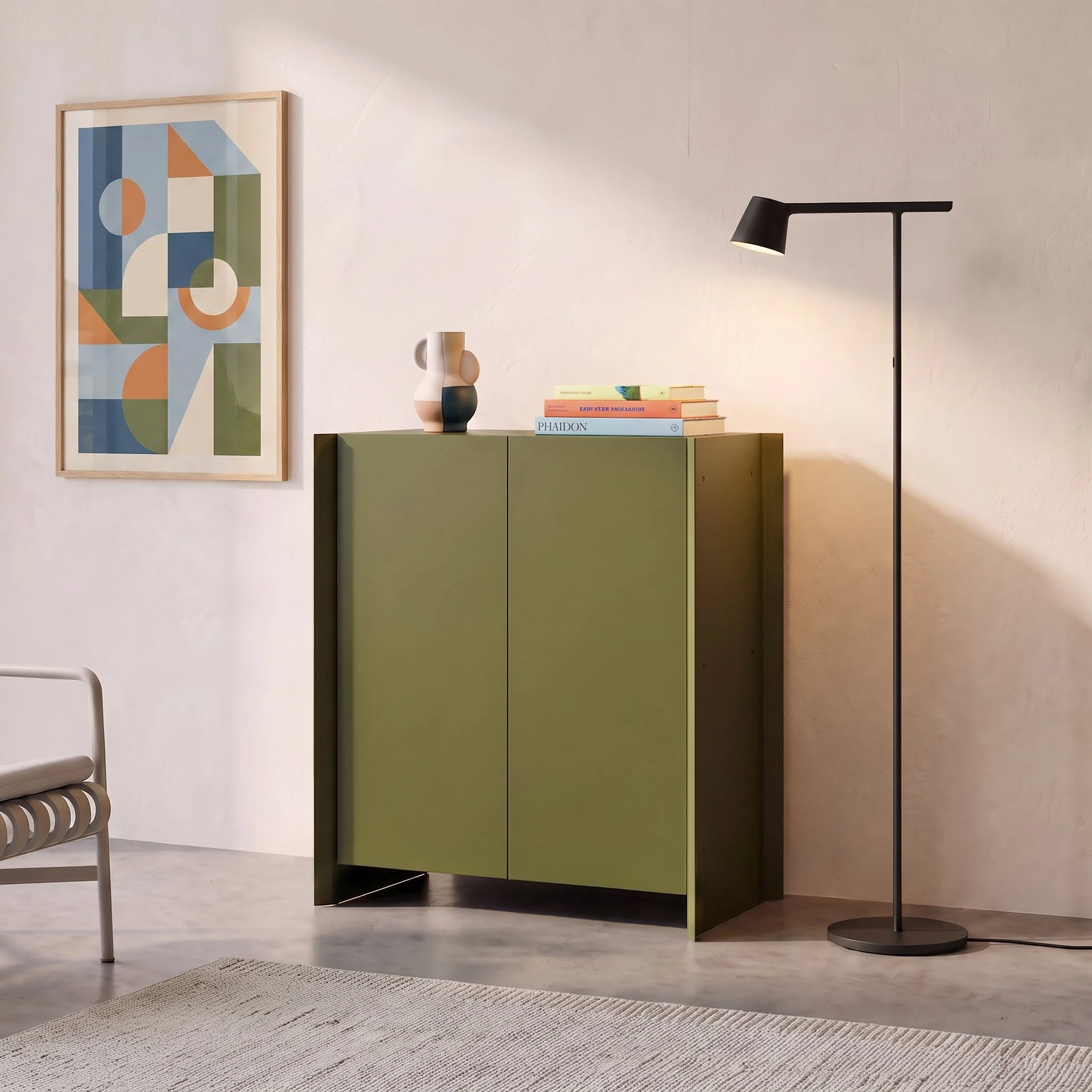 A modern minimalist interior with a colorful abstract framed art on the white wall, a green storage cabinet, decorative vases, a stack of books, a black floor lamp, and a white chair with a striped cushion.