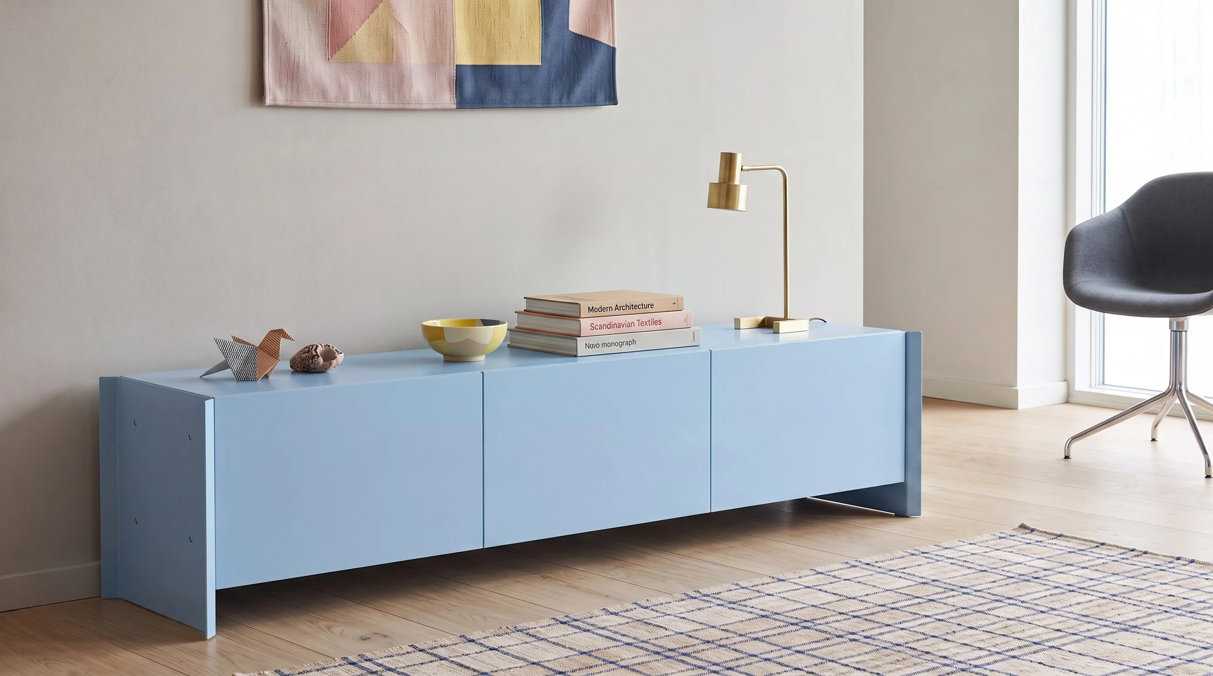 A modern interior with a pastel blue sideboard holding decorative items and books, a gold desk lamp, and a black and white geometric paper sculpture. There is a colorful, abstract wall hanging above the sideboard, a gray swivel chair near a large win