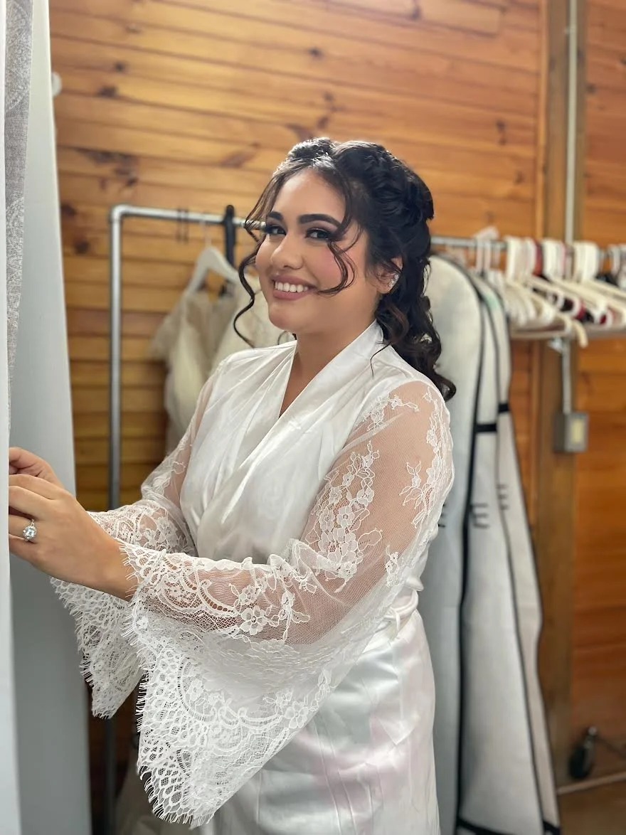 A woman in a satin robe with lace sleeves, smiling and getting ready, with a wooden wall and clothes rack in the background.