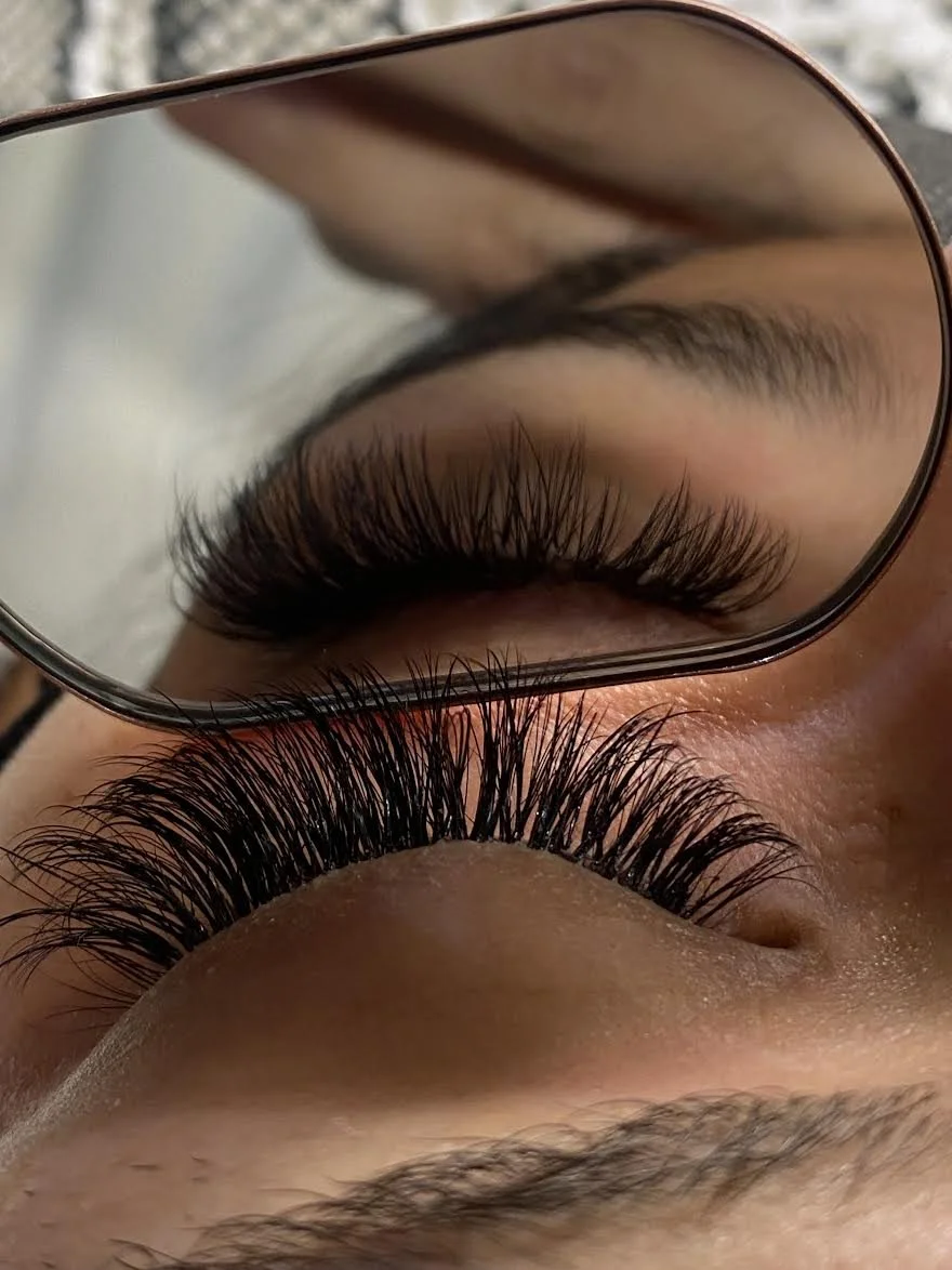 Close-up of a person's closed eye with long, curled eyelashes, viewed through a mirror.