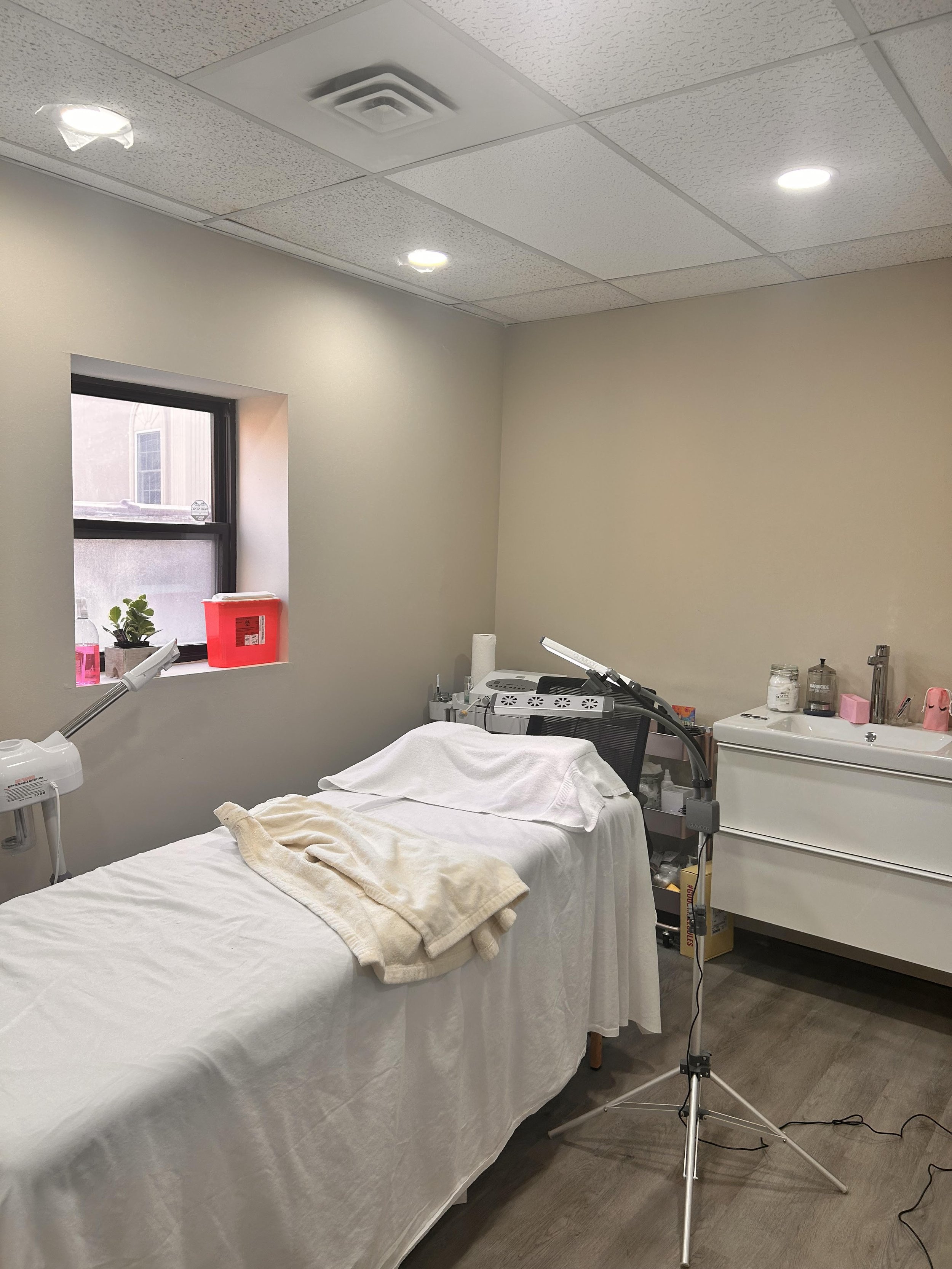 A medical or spa treatment room with a bed covered with white sheets, a folded towel, a lamp, medical equipment, cabinets, and a window, suggesting a clinical or spa setting.