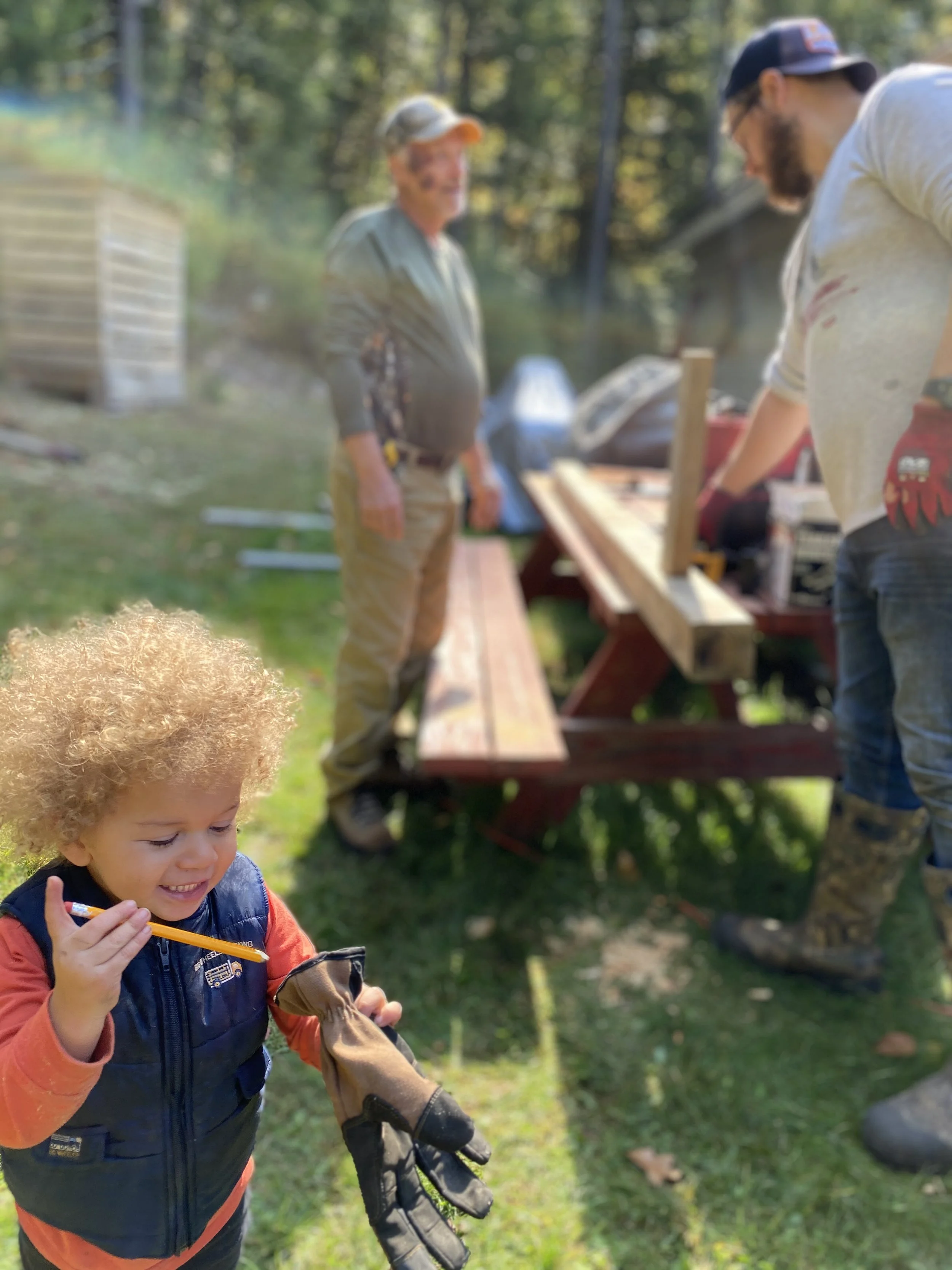 Young boy with curly blond hair smiling and holding a pair of gloves and a yellow pen, with people working on a woodworking project in the background outdoors.