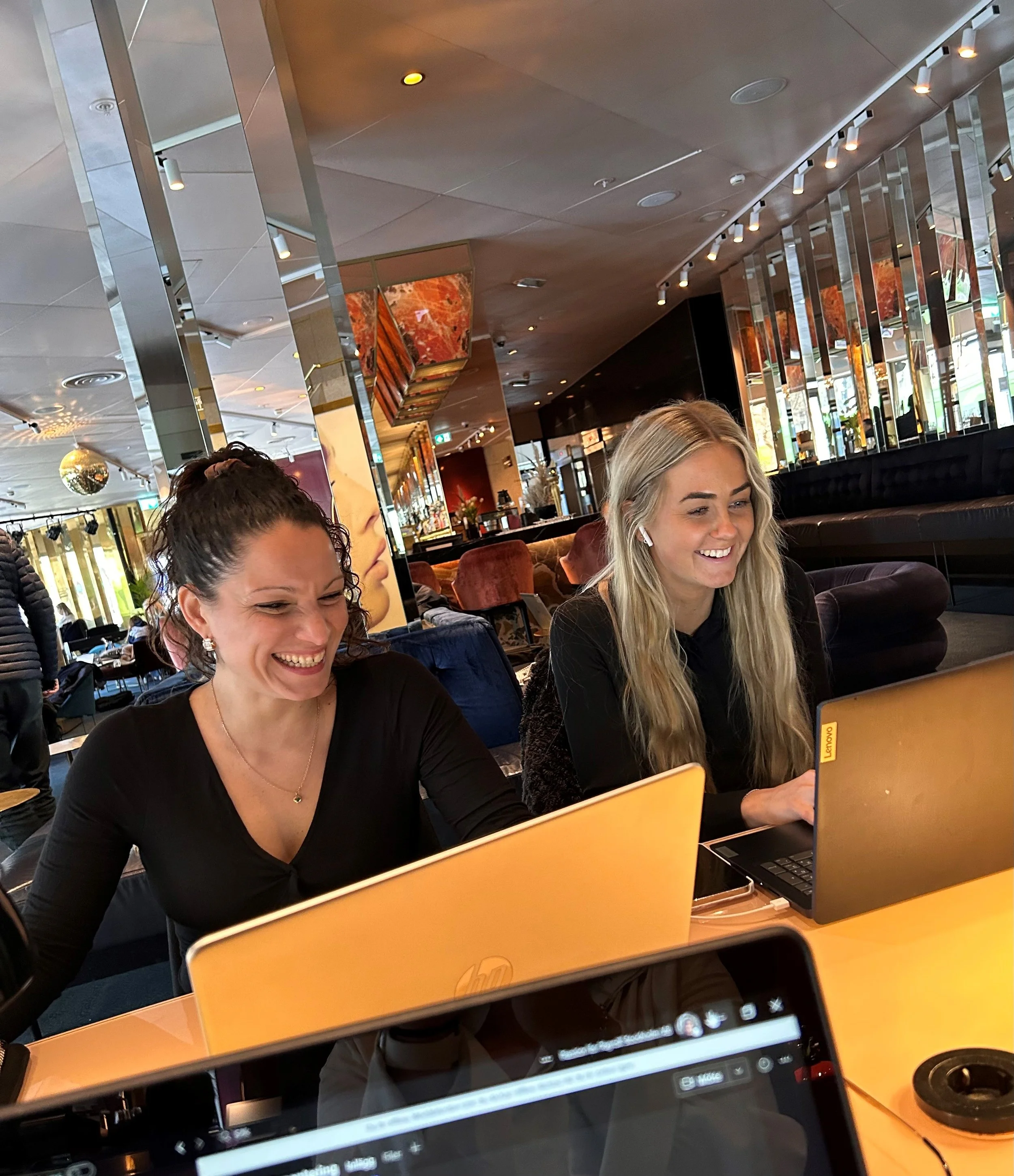 Två personer sitter vid bärbara datorer på ett modernt kontor. De ler och ser fokuserade ut. I bakgrunden syns en stilren inredning med speglar och ljus.