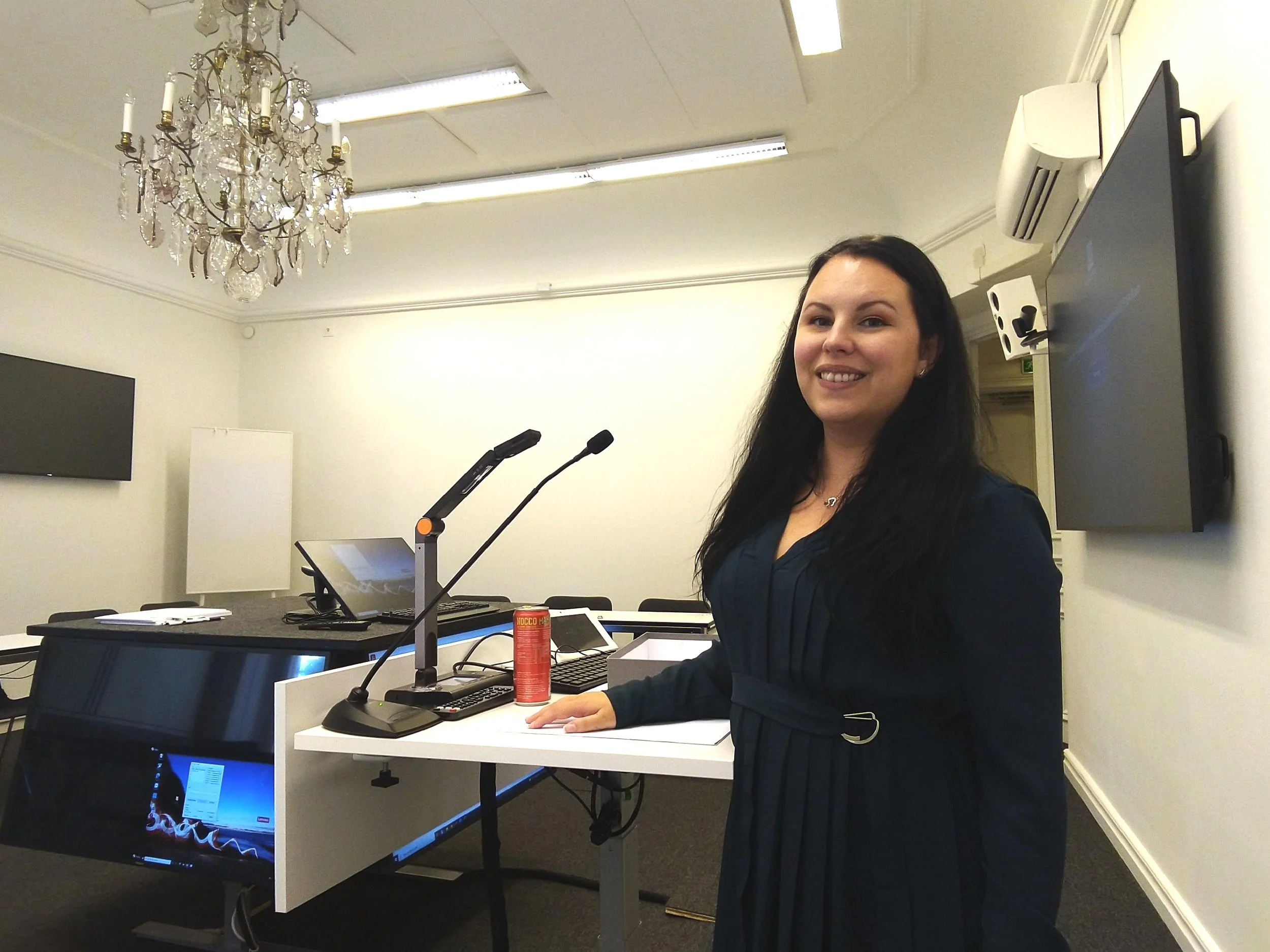 En kvinna står leende vid ett talarbord utrustat med mikrofoner och en bärbar dator i ett konferensrum. I bakgrunden syns en ljuskrona i taket och flera skärmar på väggarna.