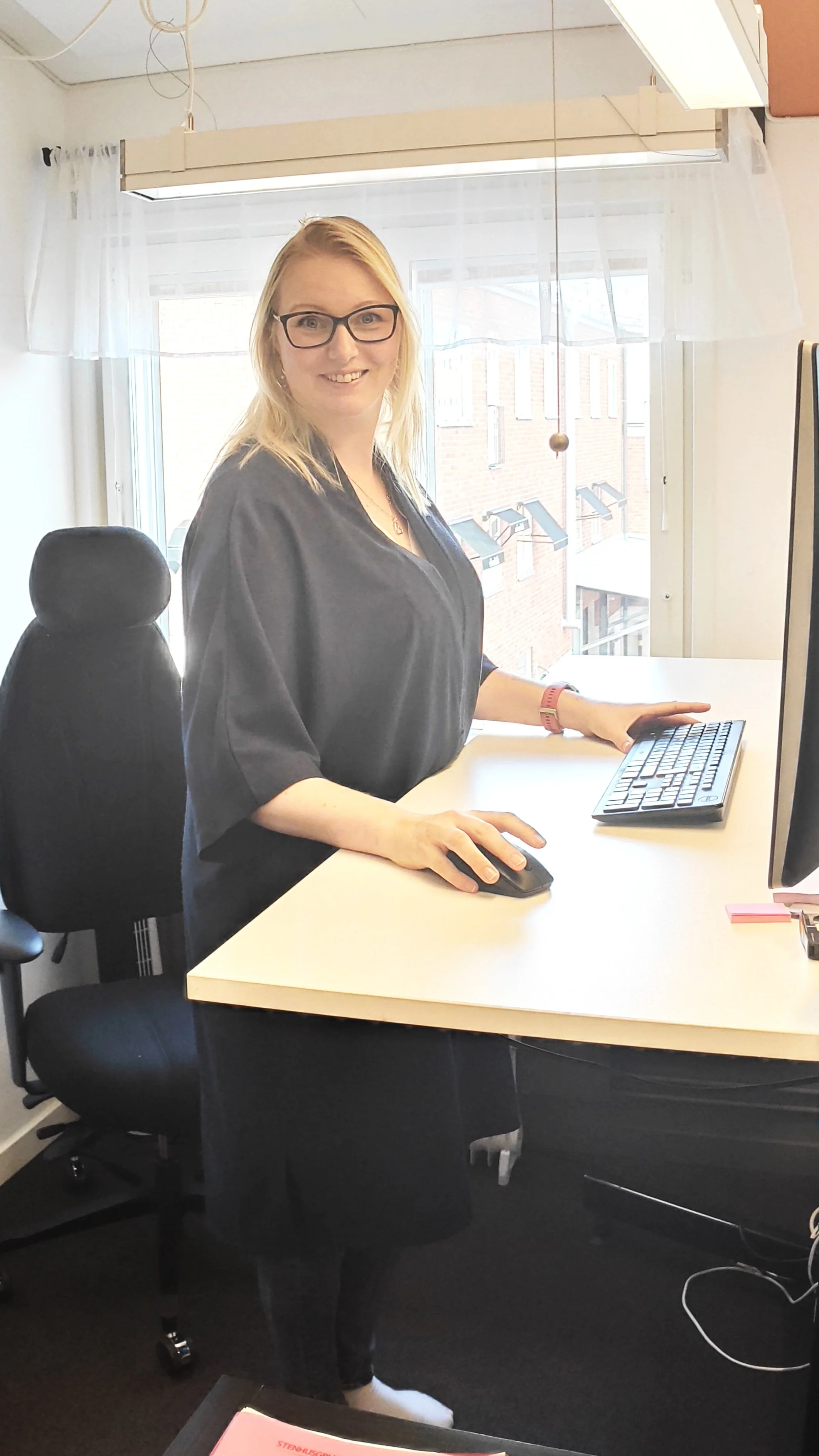 Person vid stående skrivbord med datortillbehör i ett kontorsrum.