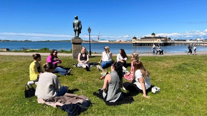 Grupp av människor som sitter i en cirkel på gräset nära vattnet, med en staty i närheten och en hamn i bakgrunden, under en klarblå himmel.