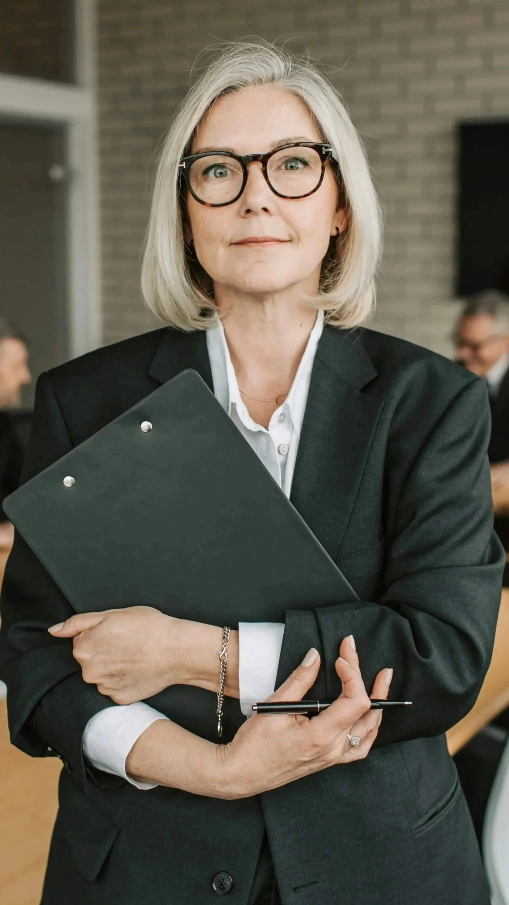 A professional woman with gray hair, wearing glasses, a dark suit, and holding a black clipboard and pen, standing in a conference room.