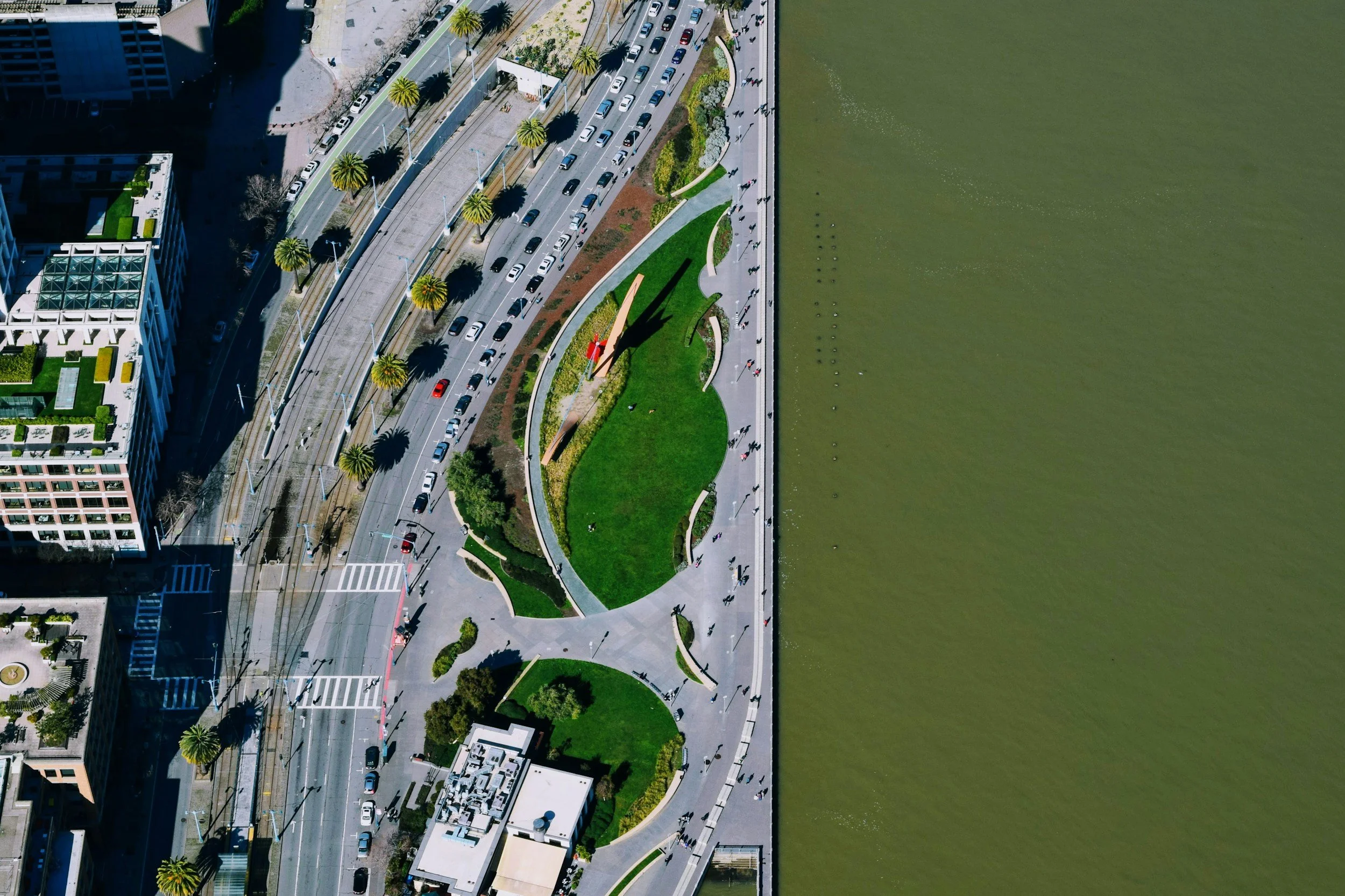 Aerial view of a city waterfront park with green lawns, walking paths, and palm trees, adjacent to a busy street with cars. The river is to the right.