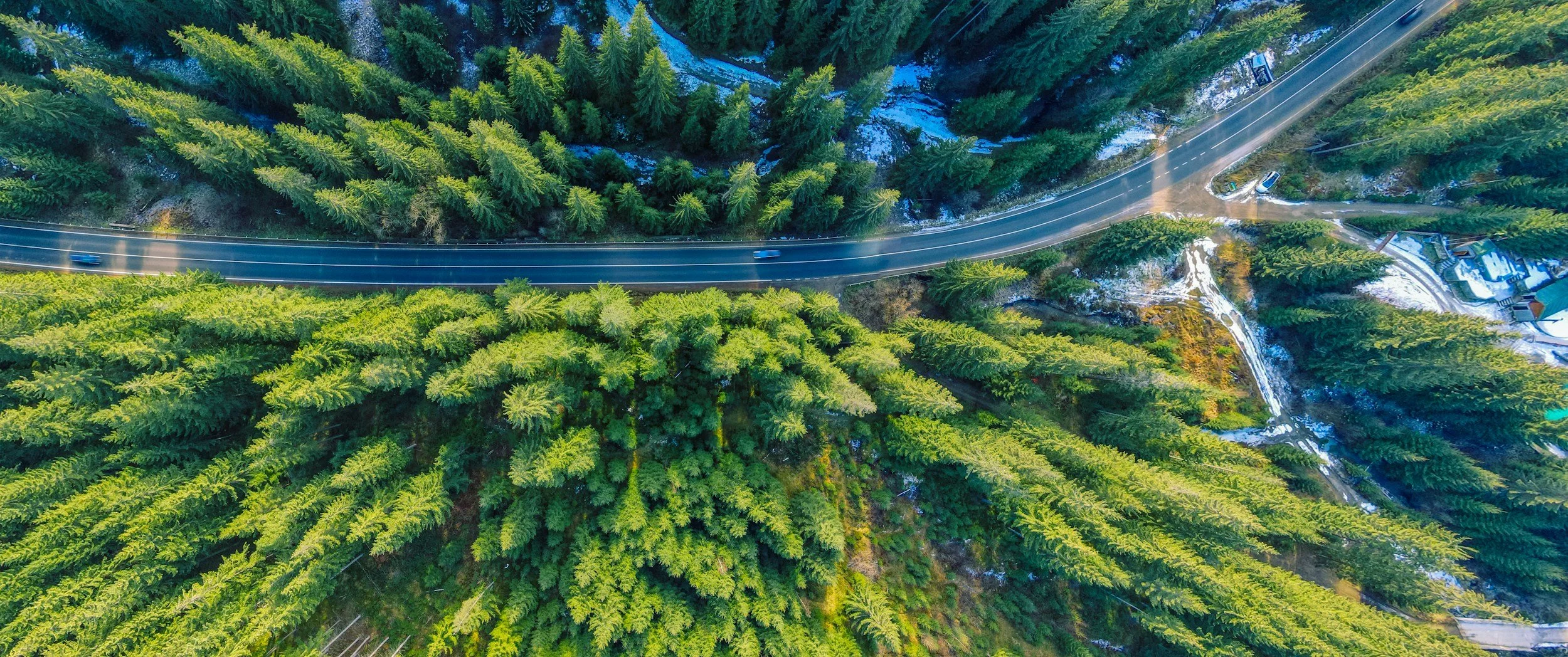 Aerial view of a winding mountain road through a dense forest with some snow patches.