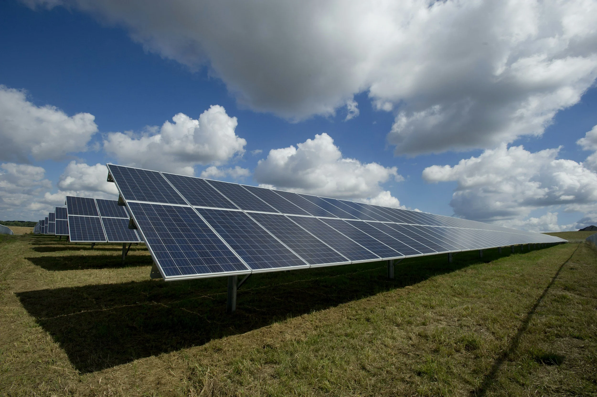 Multiple rows of solar panels installed in a grassy field under a partly cloudy sky.