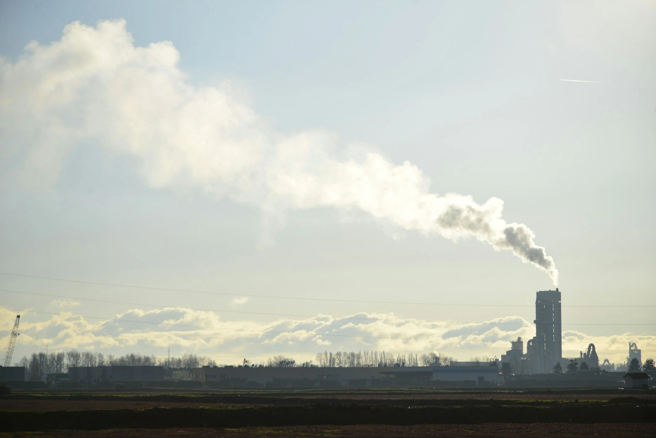 Industrial factory with smoke stacks emitting smoke into the sky, with a flat landscape and cloudy sky overhead.