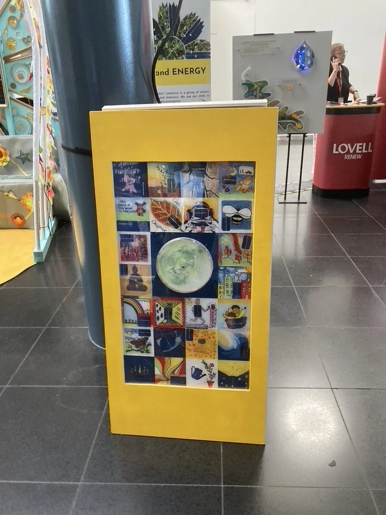  A community-created solar artwork with 24 small squares each painted by a different person to show a different perspective of energy. In the centre a larger square holds a round panel painted as the moon with moths on clock hands. Each small square 