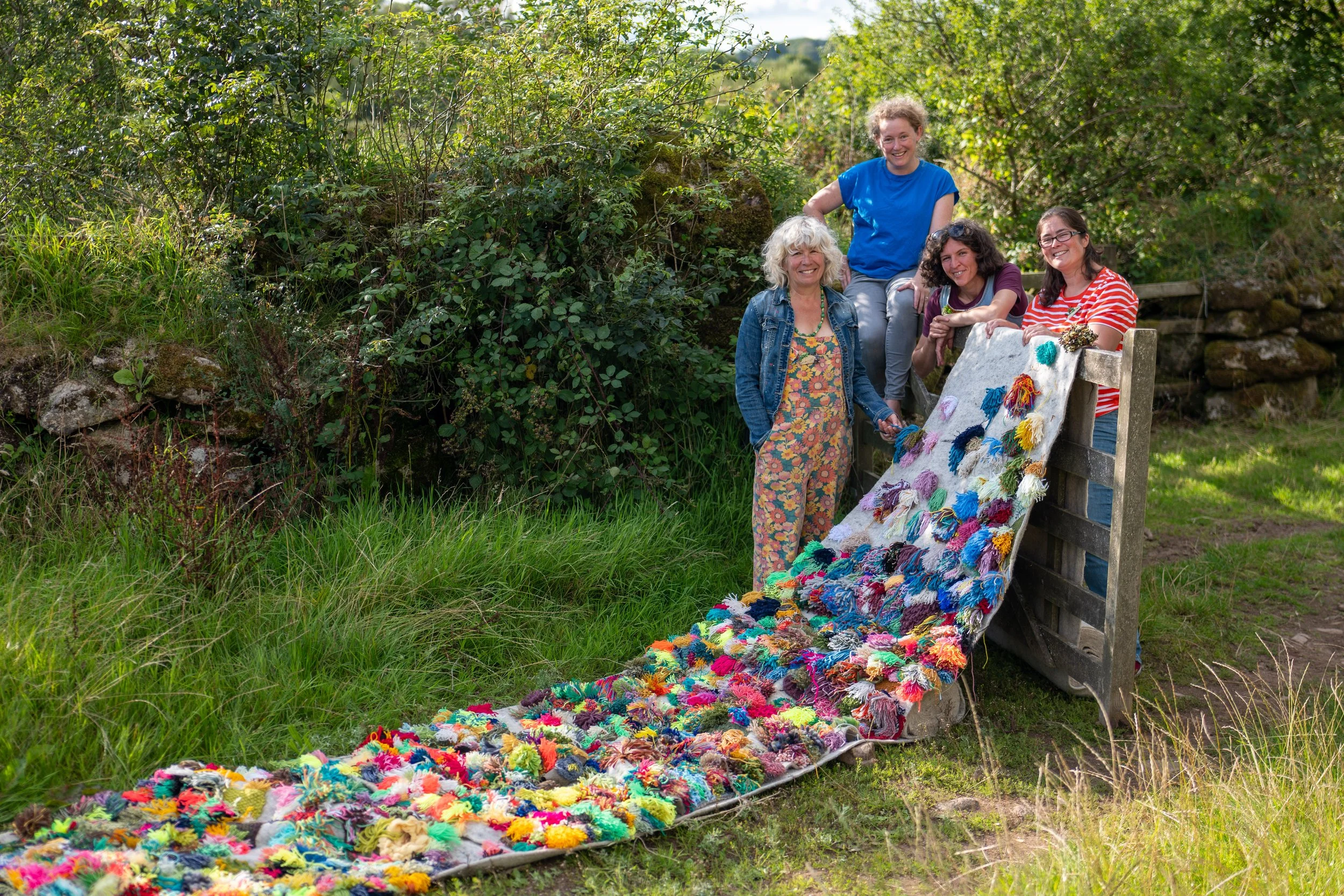 Four people standing outdoors next to a colorful textile art piece hanging from a wooden structure, surrounded by greenery.