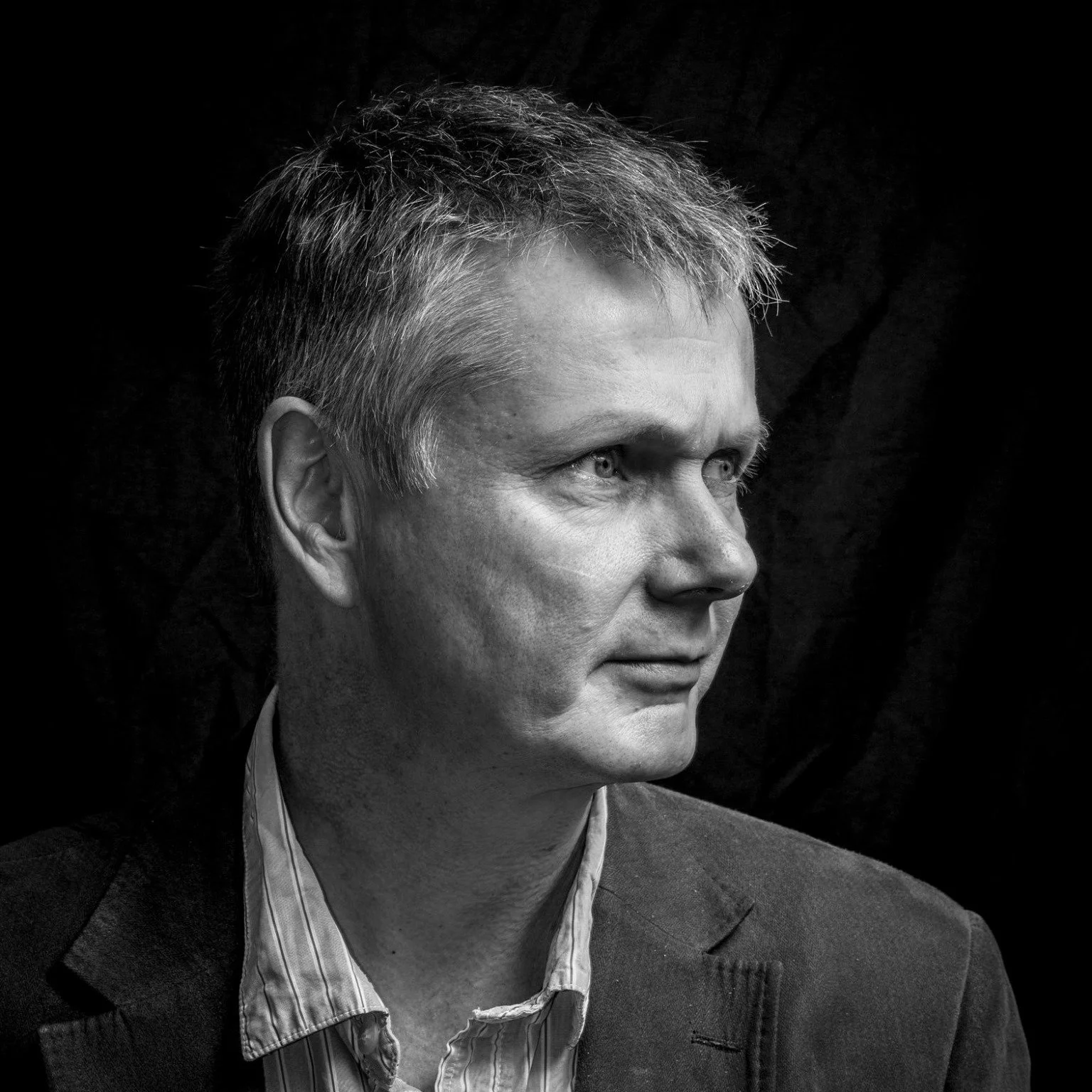 Black and white portrait of a man looking sideways, wearing a suit jacket and a striped shirt, against a dark background.