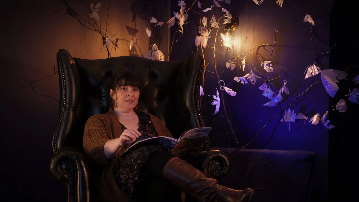 Woman sitting in a large leather chair, holding a book, surrounded by branches with paper butterflies, warmly lit.