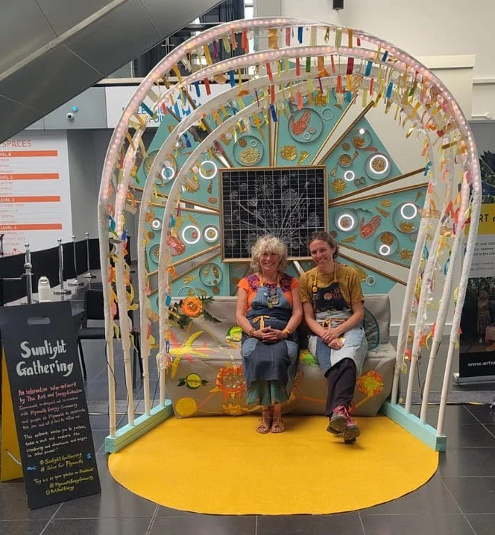 Naomi and Jenny sit on the bench of Sunlight Gathering - a large vibrant interactive solar powered artwork