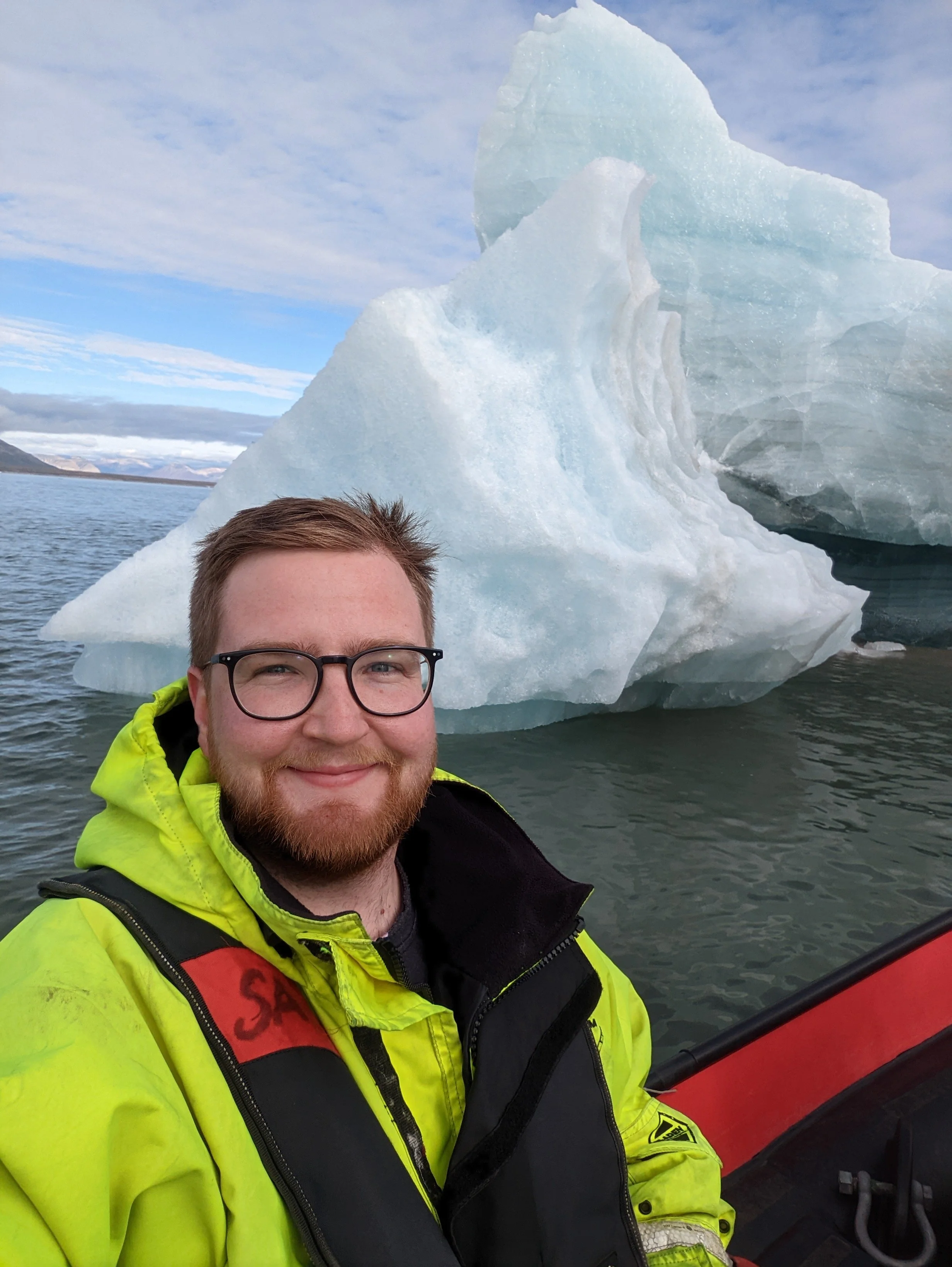 Person in a yellow jacket smiling in front of an iceberg on a cloudy day.