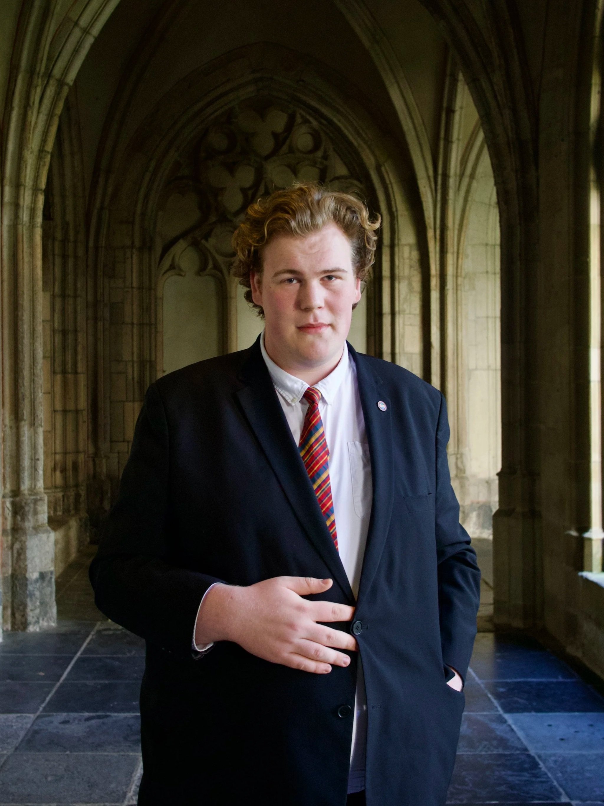 Jongen in formeel zwart pak met gekleurde stropdas in een historische kerk of kasteel, met decoratieve gewelven op de achtergrond.